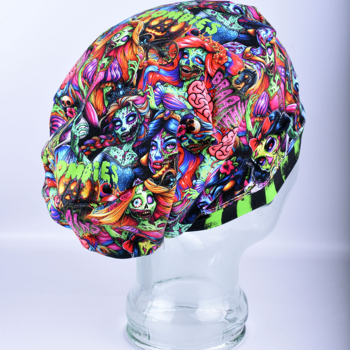 A multicolor skull-shaped fabric scrub cap featuring a vibrant, detailed graphic print of skulls, zombies, and brains, displayed on a clear glass mannequin head against a light gray background.