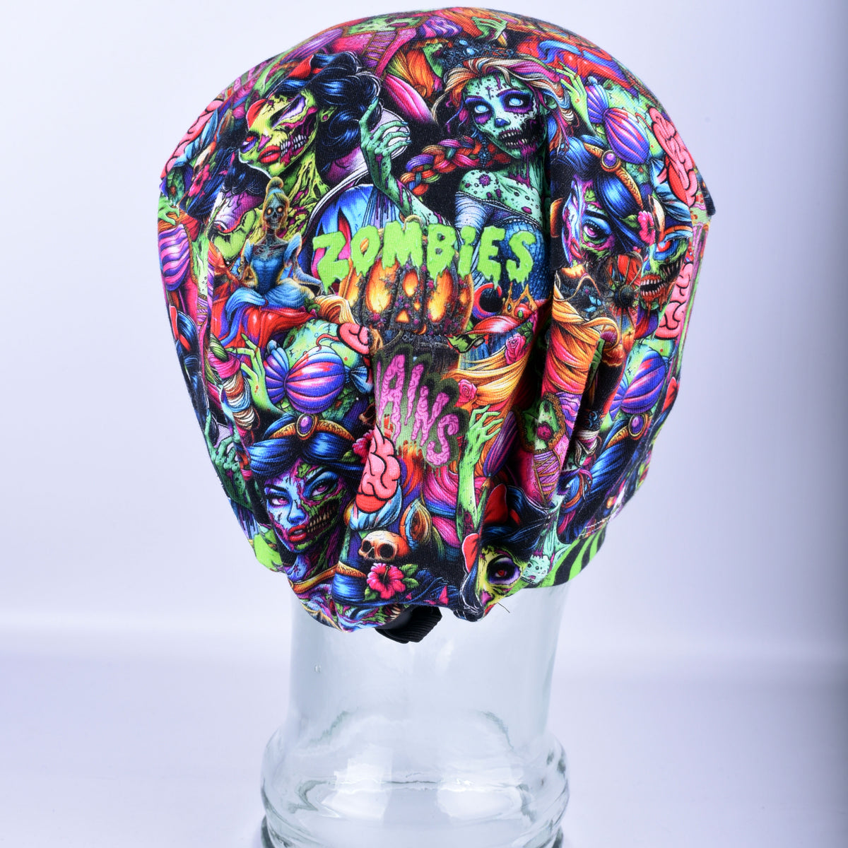 A multicolor Halloween-themed scrub cap featuring a vibrant zombie and horror graphic print, displayed on a clear glass mannequin head against a light gray background.