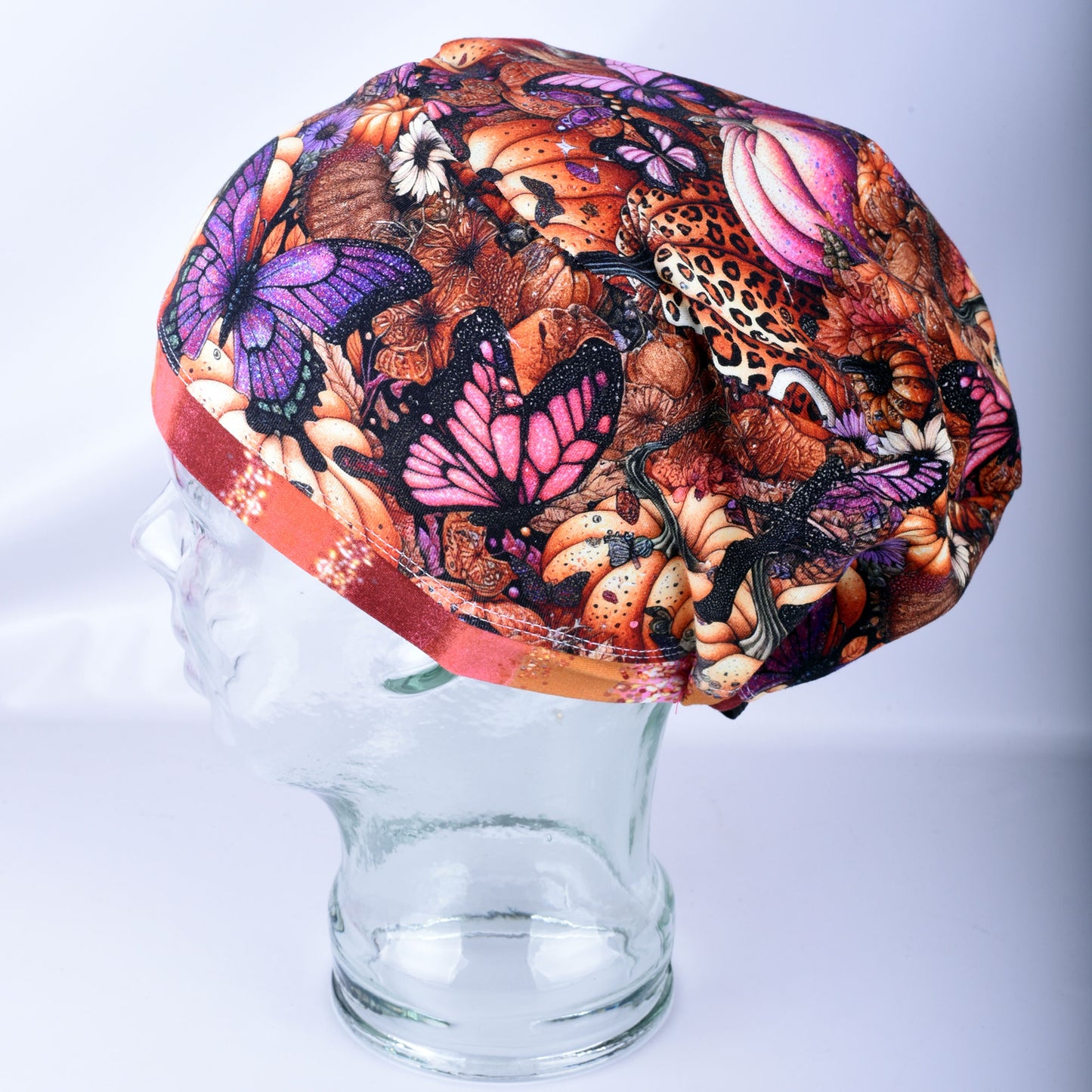 A multicolor fabric surgical scrub cap featuring a busy pattern of purple, pink, orange, and brown butterflies, flowers, pumpkins and leaves, displayed on a clear glass mannequin head against a light gray background.