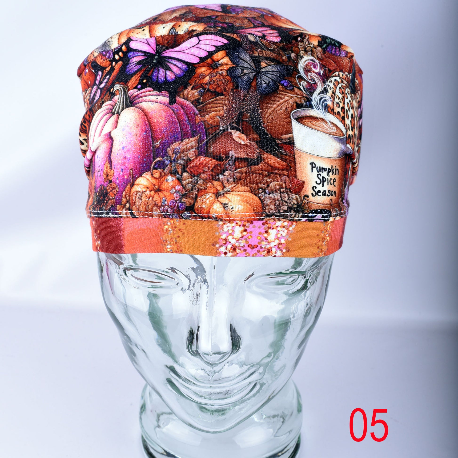 A multicolor autumn-themed fabric scrub cap displayed on a clear glass mannequin head. The pattern features pumpkins, butterflies, leaves, and a coffee cup labeled 'Pumpkin Spice Season'.