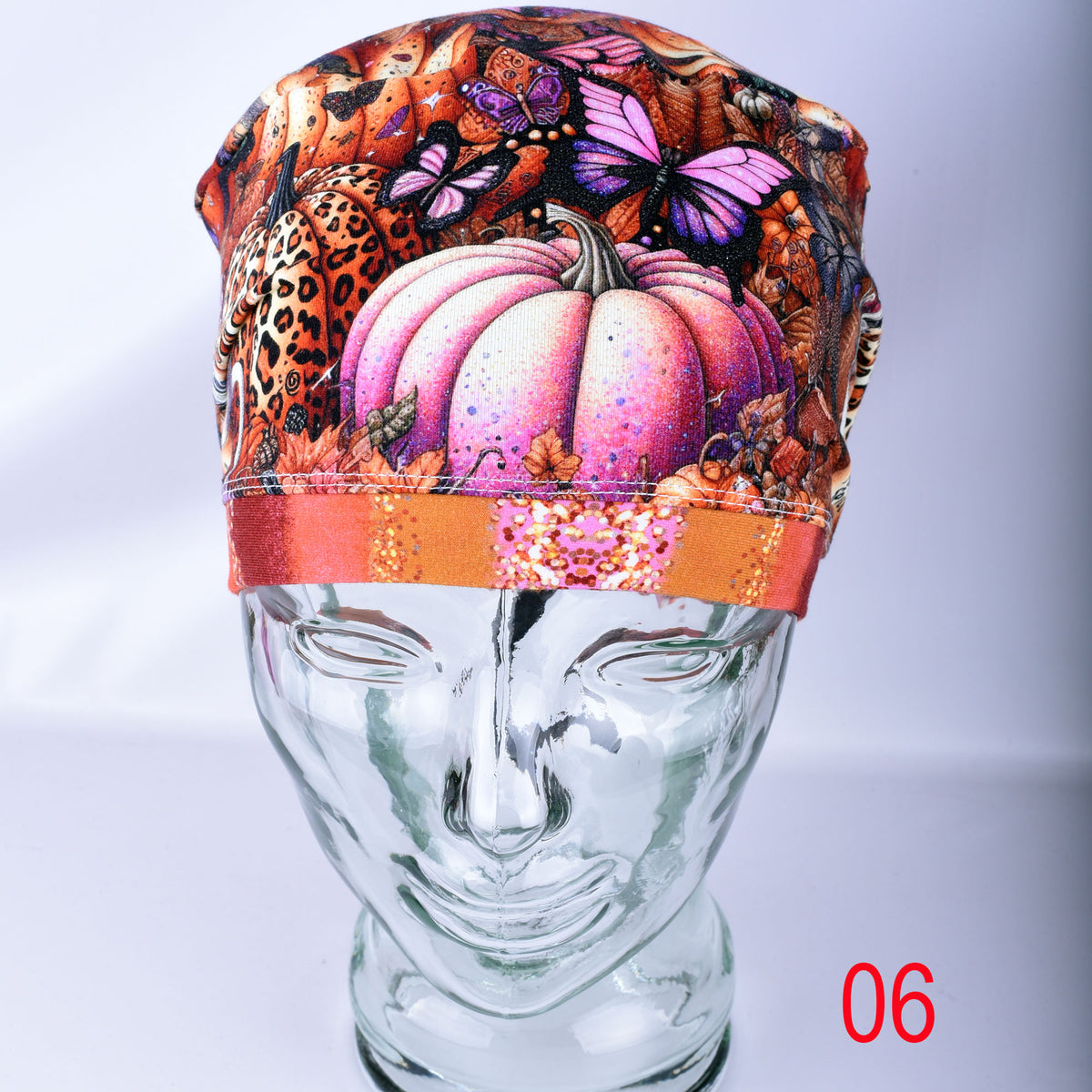 A multicolor printed surgical cap featuring pumpkins, butterflies, and leopard print, displayed on a clear glass mannequin head. The cap has a gradient orange band.