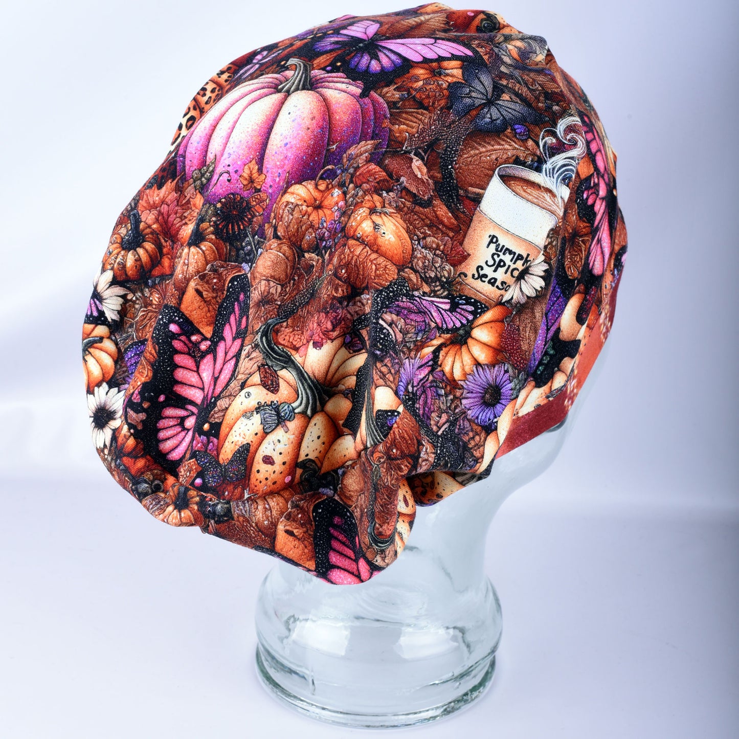 A fabric scrub cap featuring a detailed autumn pattern with pumpkins, butterflies, and a coffee cup graphic labeled 'Pumpkin Spice Season,' displayed on a clear glass mannequin head against a light gray background.