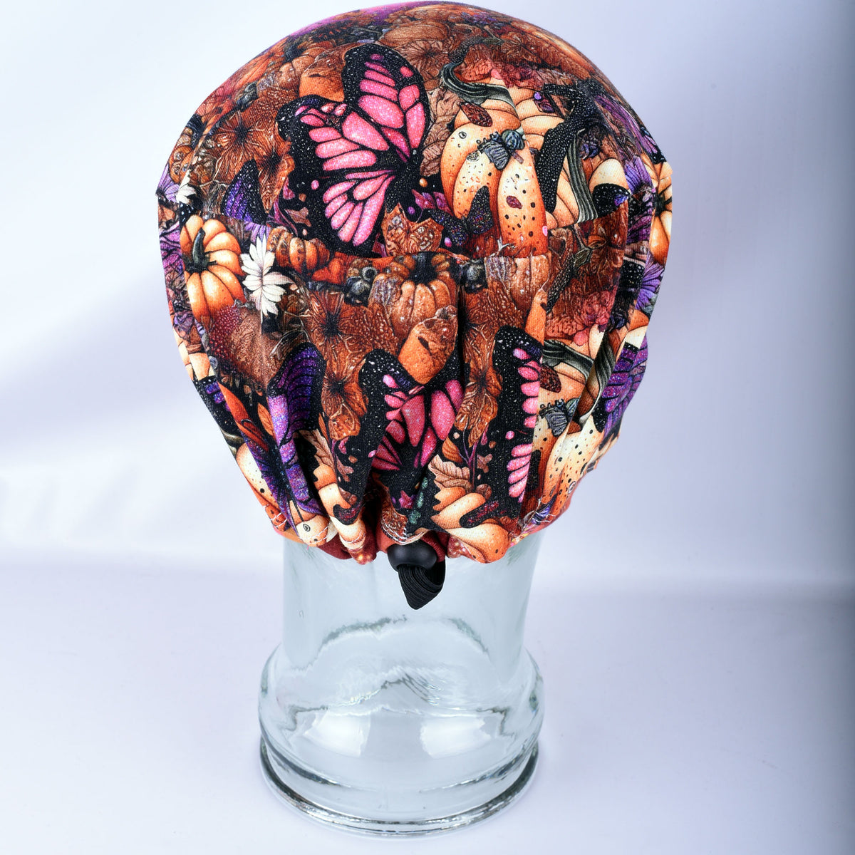 A fabric scrub cap with a busy autumn butterfly and pumpkin print, displayed on a clear glass mannequin head against a light gray background.