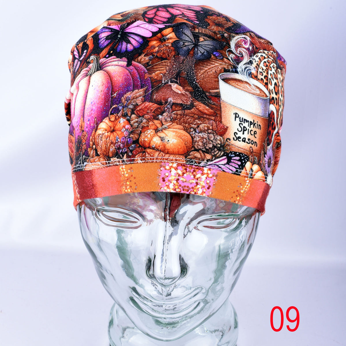 A patterned scrub cap featuring an autumn theme with pumpkins, butterflies, and a coffee cup graphic reading 'Pumpkin Spice Season,' displayed on a clear mannequin head. A red '09' text overlay is visible in the bottom right corner.