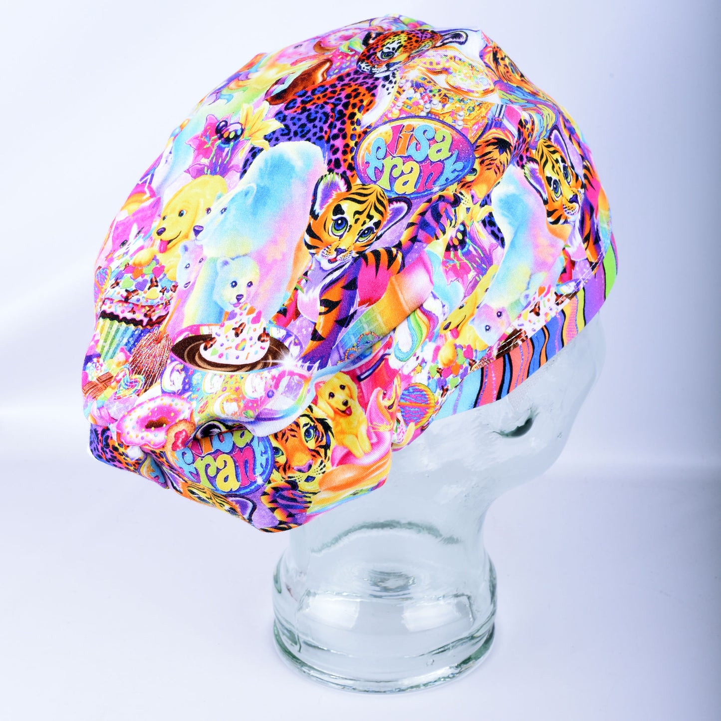 A multicolor, highly patterned fabric scrub cap displayed on a clear glass mannequin head against a light gray studio background. The busy design features cartoon tigers, cupcakes, planets, and colorful abstract shapes.