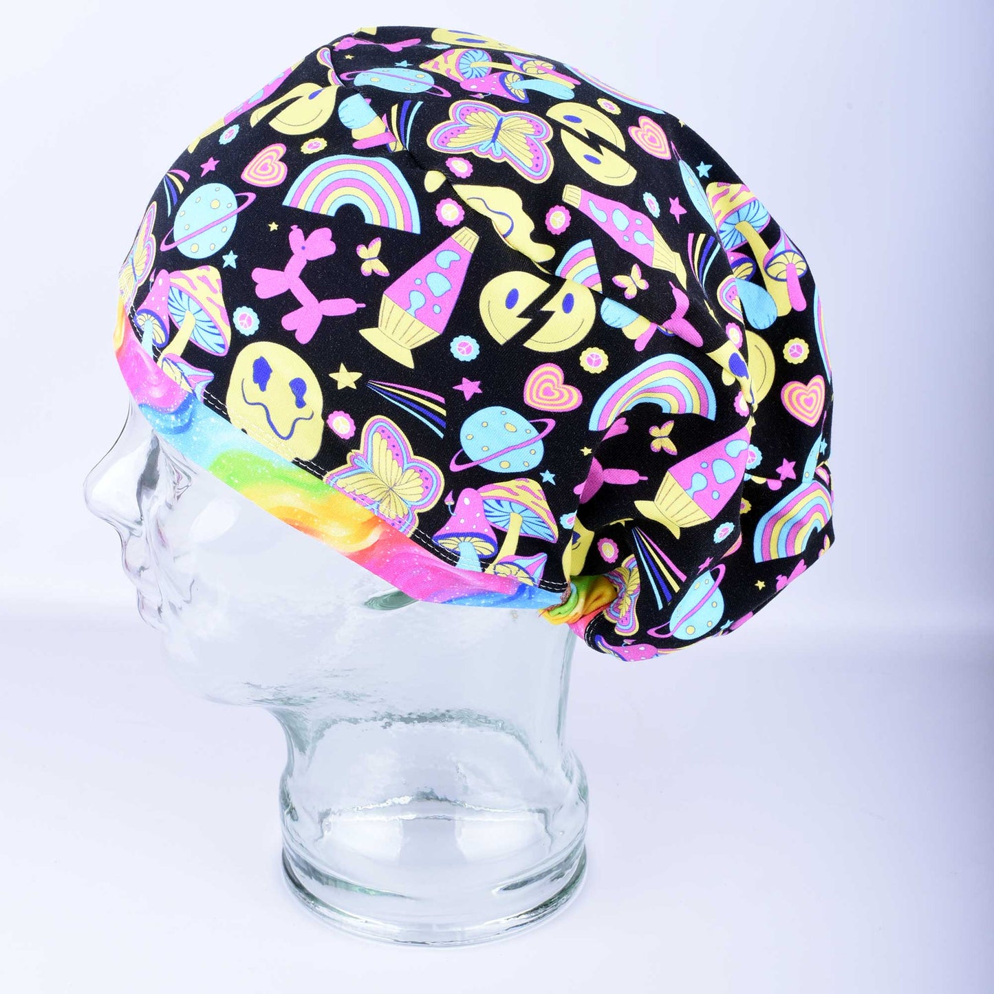 A retro days style full coverage scrub cap displayed on a clear mannequin head against a light blue-gray background. The medical hat features a vibrant, colorful black fabric patterned with smiley faces, rainbows, mushrooms, peace signs, and butterflies, accented with a solid pink brim.