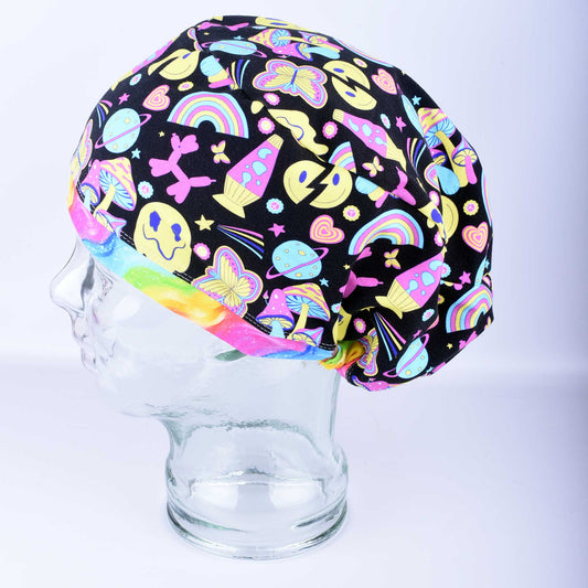 A retro days style full coverage scrub cap displayed on a clear mannequin head against a light blue-gray background. The medical hat features a vibrant, colorful black fabric patterned with smiley faces, rainbows, mushrooms, peace signs, and butterflies, accented with a solid pink brim.