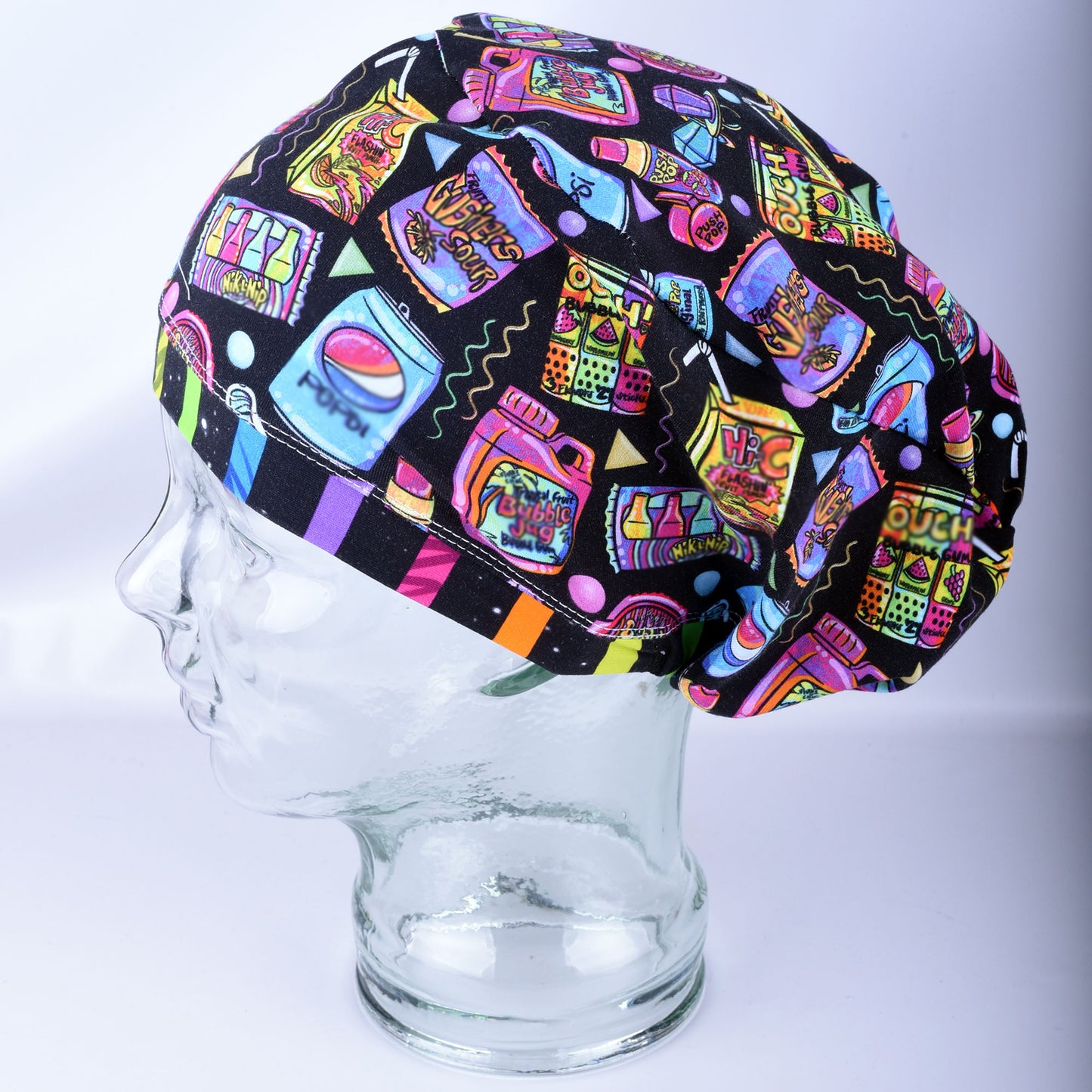 A multicolor retro-themed scrub cap featuring a vibrant pop-art pattern of candy, bubblegum, and soda cans on a black background, displayed on a clear glass mannequin head.