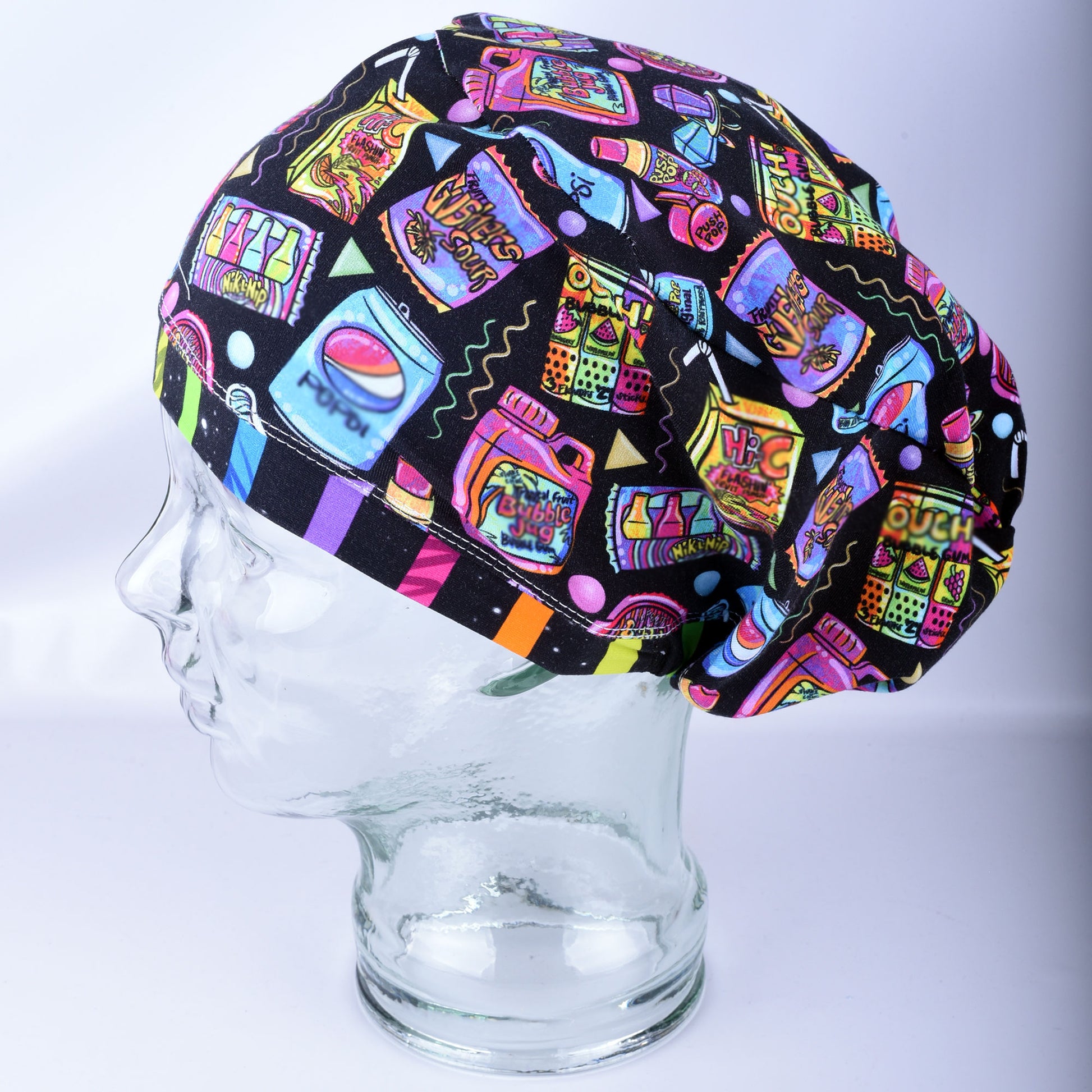 A multicolor retro-themed scrub cap featuring a vibrant pop-art pattern of candy, bubblegum, and soda cans on a black background, displayed on a clear glass mannequin head.