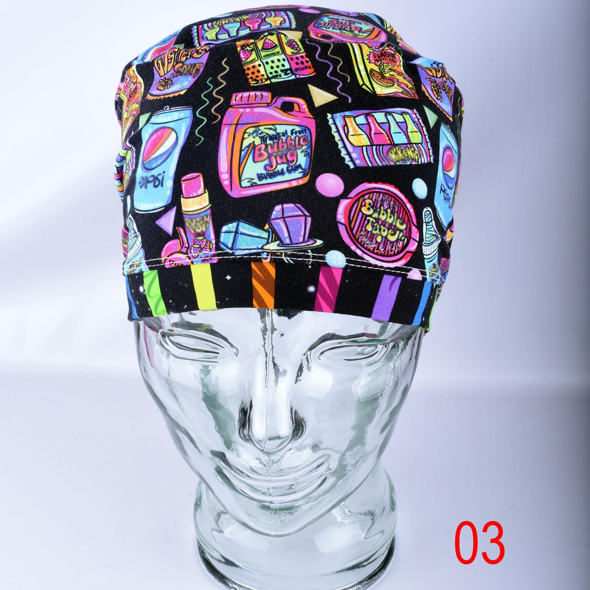 A black surgical-style cap with a colorful retro pop-art print, displayed on a clear glass mannequin head. The print features neon pink, yellow, blue, and green graphics including a soda can, bubblegum, and candy.