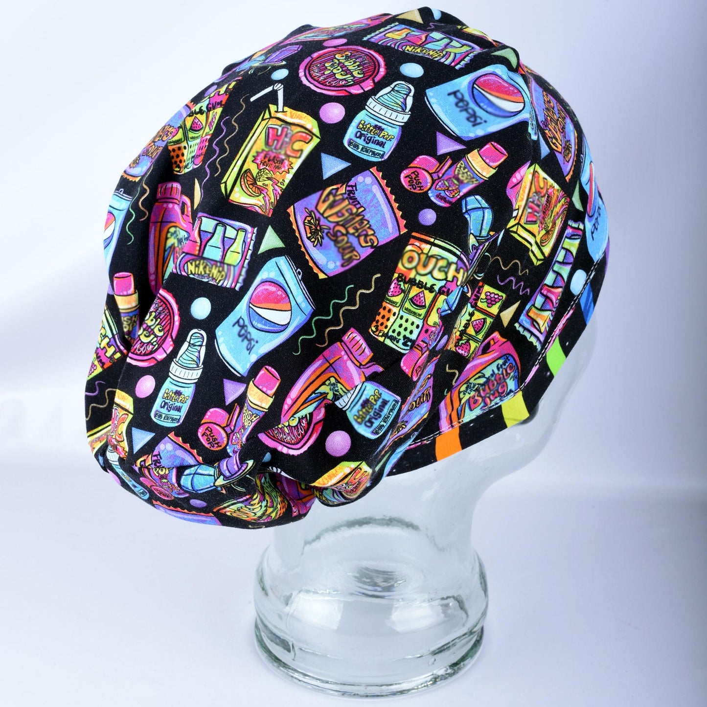 A black scrub cap featuring a colorful, vibrant graphic print of various candy, bubblegum, and soda pop illustrations, displayed on a clear glass mannequin head against a light gray background.