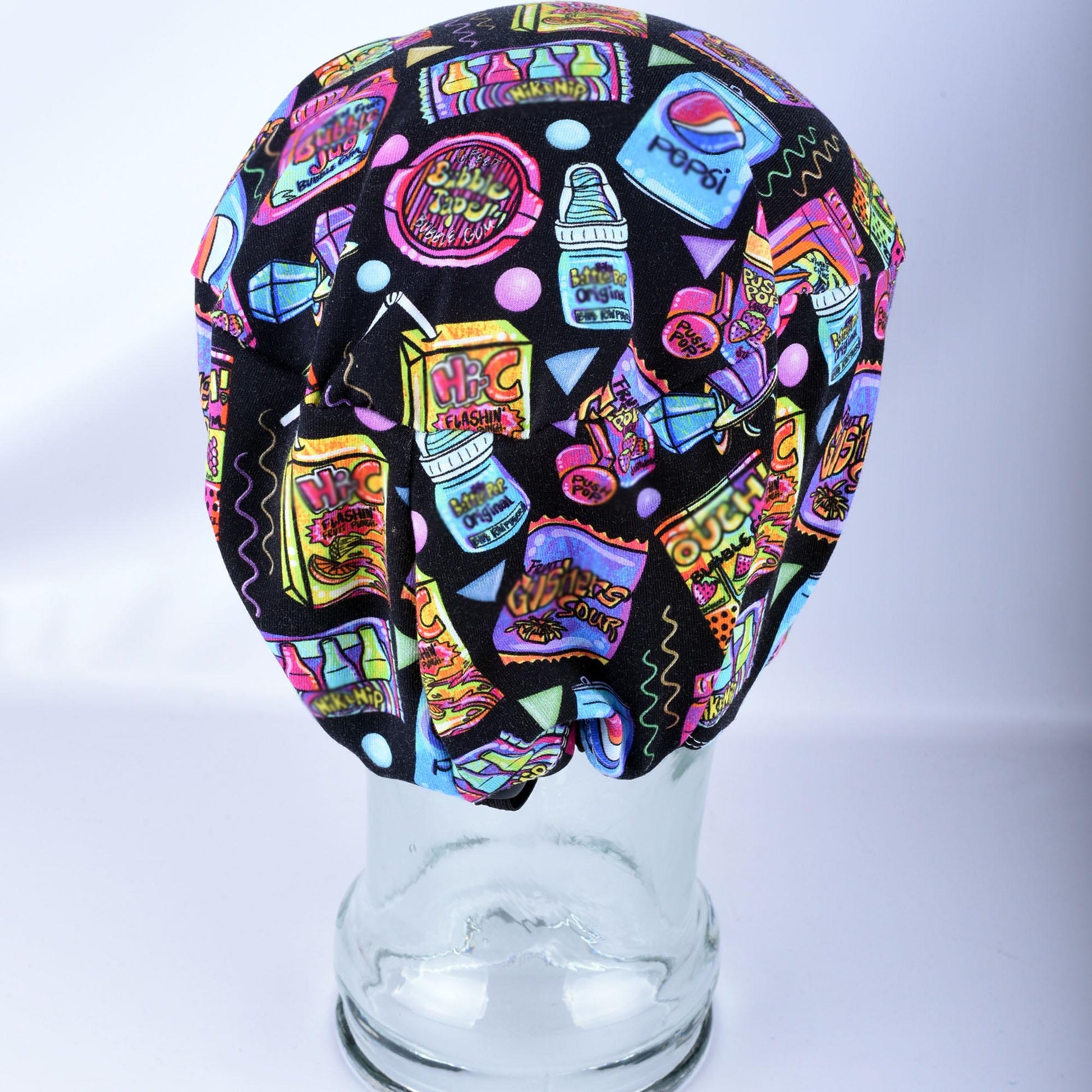 Back view of a black scrub cap featuring a colorful retro graphic print of various soda bottles and candy, displayed on a clear glass mannequin head against a white background.