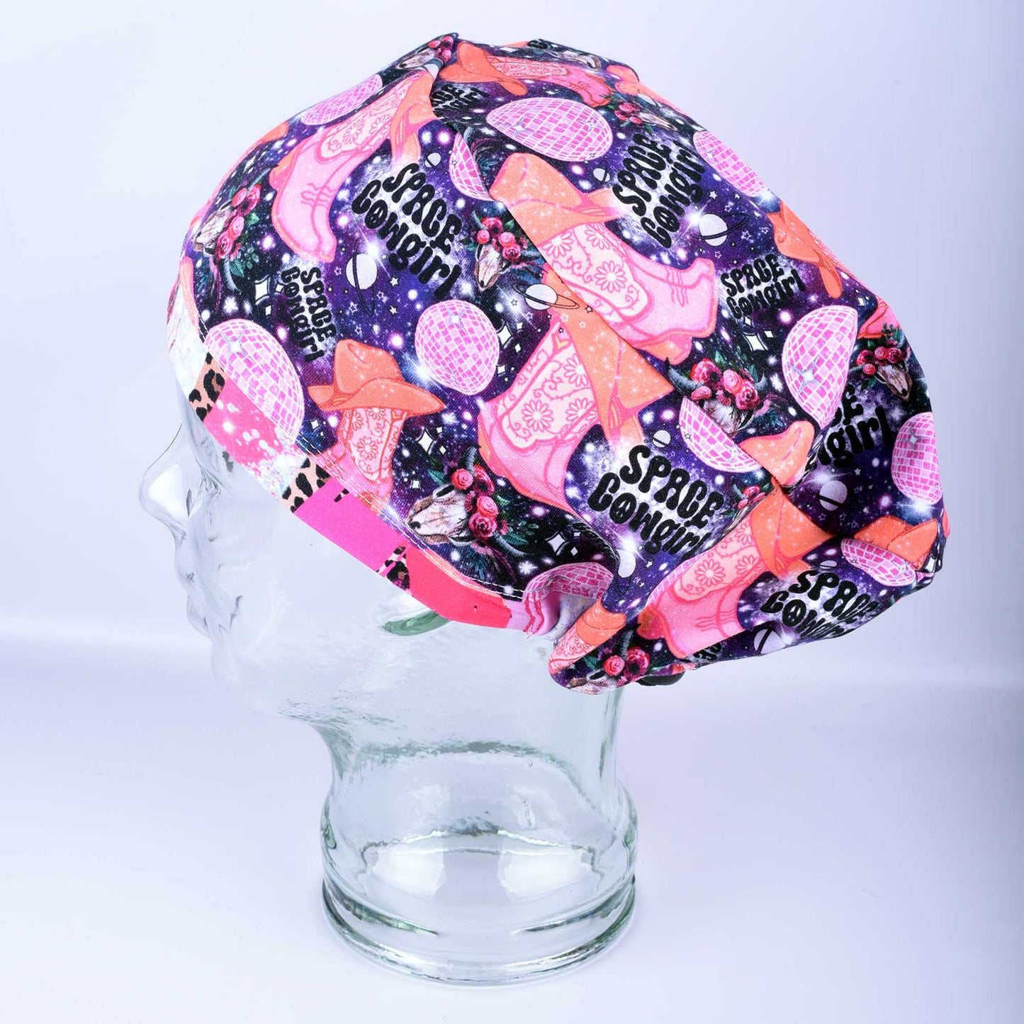Close-up of the Space Cowgirl scrub cap fabric featuring a vibrant pattern of disco balls, cowboy boots, stars, and crescent moons on a dark background.