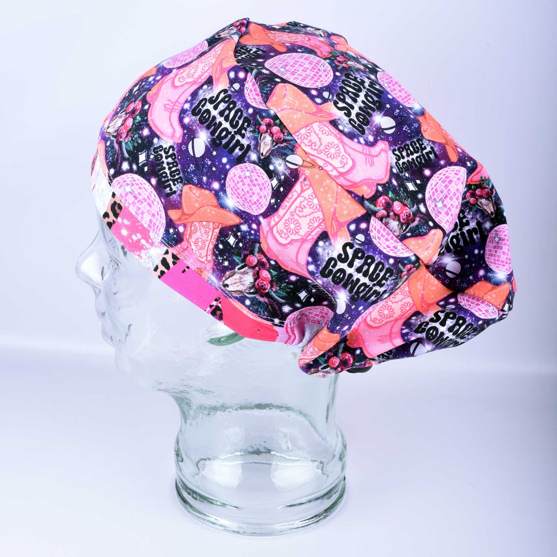 Close-up of the Space Cowgirl scrub cap fabric featuring a vibrant pattern of disco balls, cowboy boots, stars, and crescent moons on a dark background.