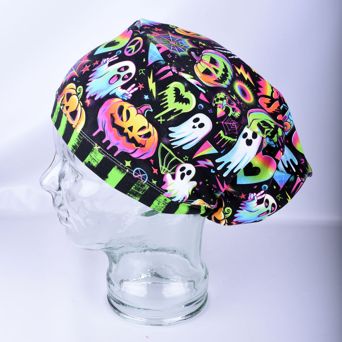 A black scrub cap with a vibrant neon Halloween print featuring ghosts, pumpkins, and lightning bolts, displayed on a clear glass mannequin head against a light gray background.