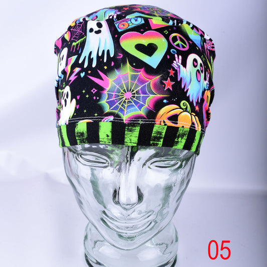 A neon Halloween-themed surgical scrub cap displayed on a clear glass mannequin head. The cap features a black background decorated with colorful, glowing graphics including ghosts, a heart, a spiderweb, and a pumpkin, finished with a striped black and neon green band.