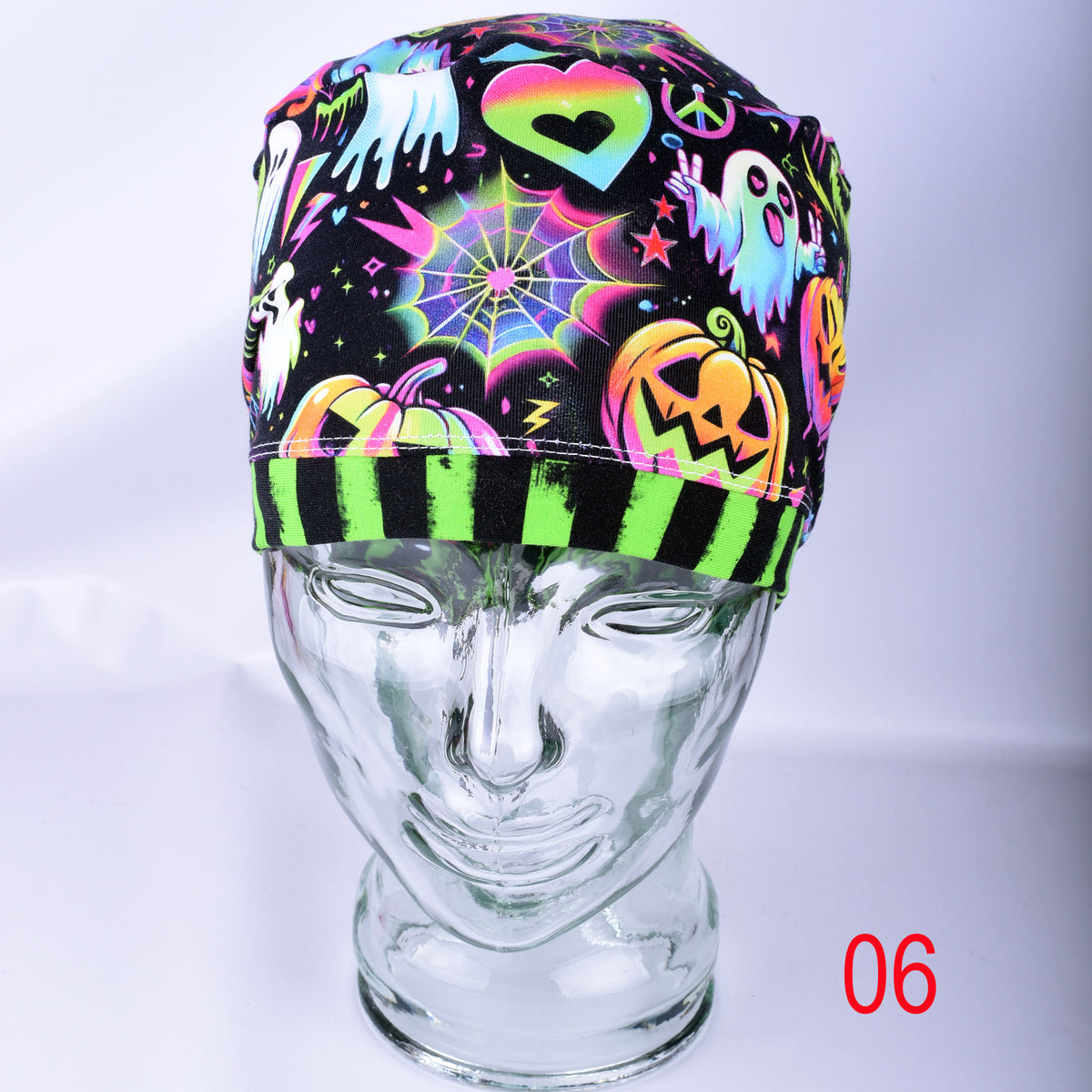 A neon Halloween-themed surgical scrub cap displayed on a clear glass mannequin head. The cap features a black background decorated with glowing pink, green, orange, and blue graphics including ghosts, pumpkins, a spiderweb, and a peace sign.