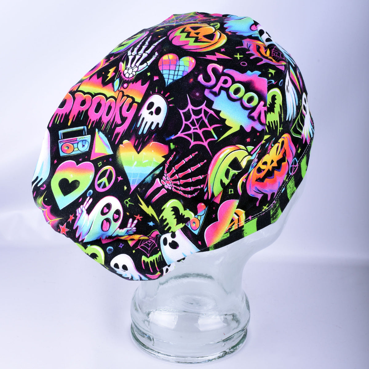 A black medical scrub cap with a vibrant, multicolored Halloween-themed graphic print, displayed on a clear glass mannequin head against a light gray background.