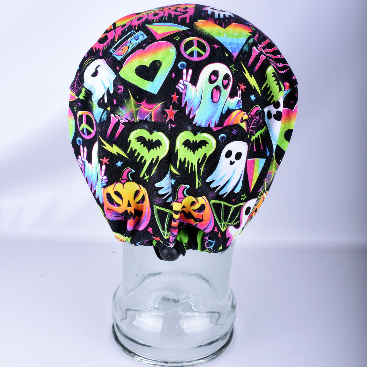Back view of a black scrub cap featuring a vibrant neon Halloween pattern with ghosts, pumpkins, hearts, and peace signs, displayed on a clear glass jar stand against a white background.