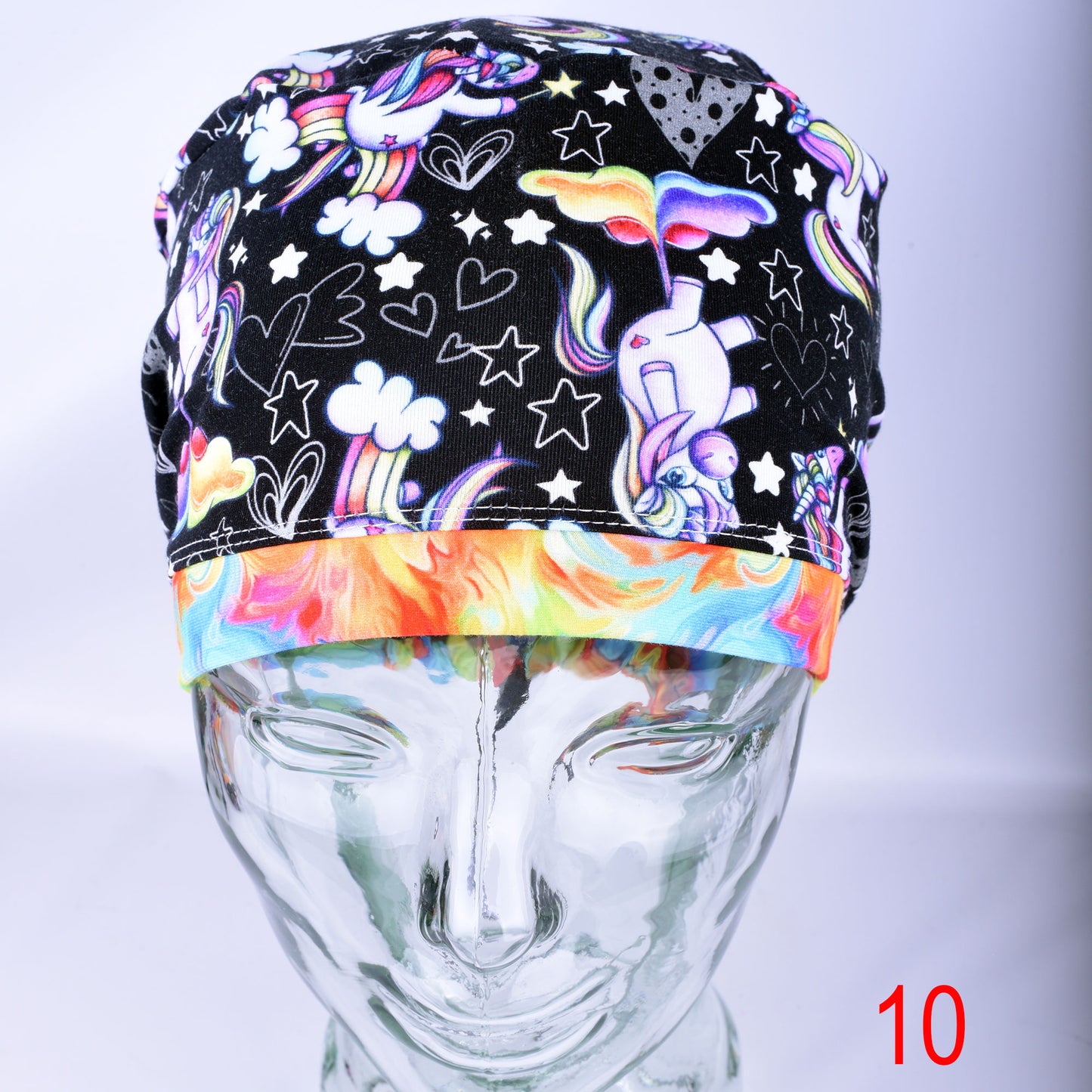 A close-up view of a fabric scrub cap on a clear mannequin head, featuring a colorful print of whimsical unicorns, stars, and rainbow hearts against a dark, patterned background.