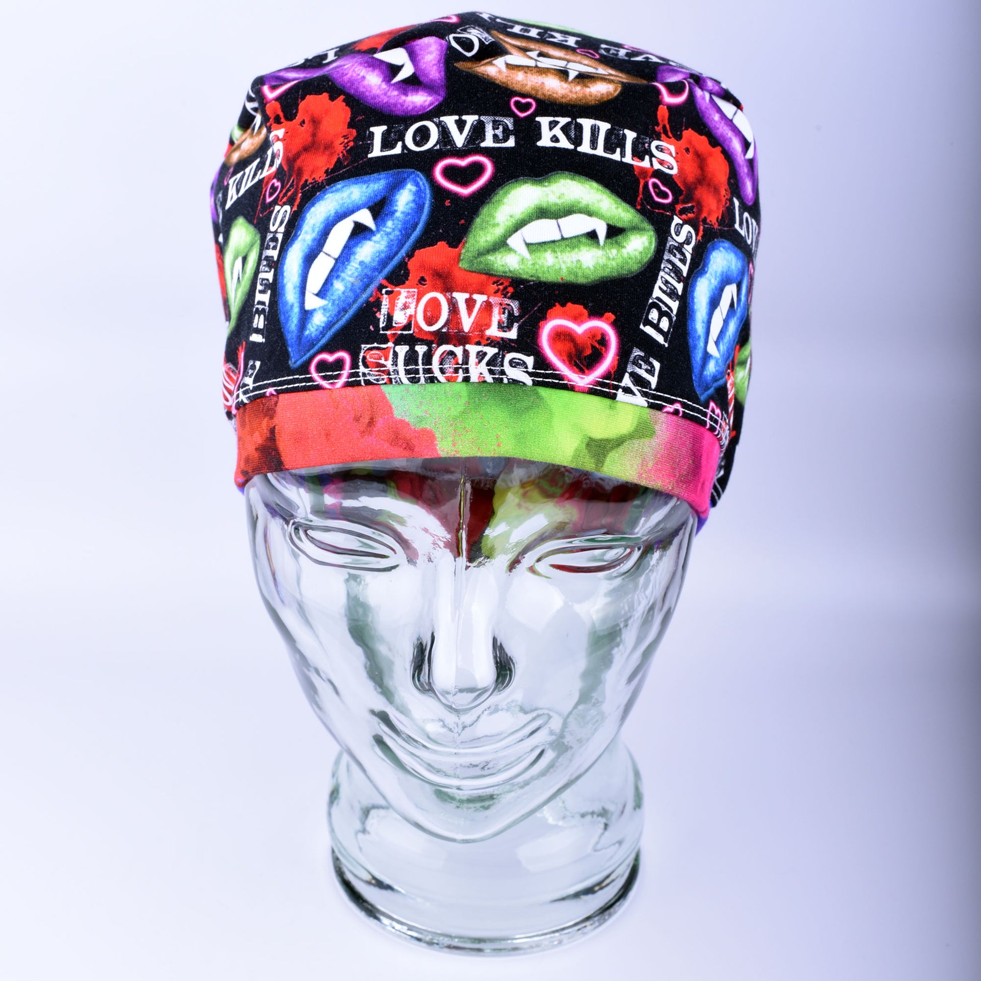 Full-coverage scrub cap featuring an edgy, vibrant Valentine's Day pattern of colorful conversation hearts with phrases like "Love Kills" and "Love Sucks" on a black background, displayed on a clear glass mannequin head.