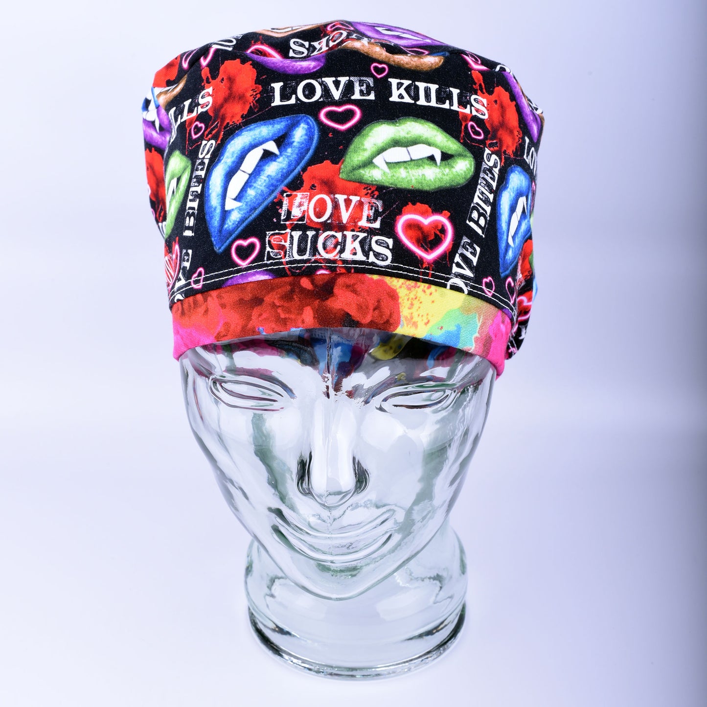Full-coverage scrub cap featuring an edgy, vibrant Valentine's Day pattern of colorful conversation hearts with phrases like "Love Kills" and "Love Sucks" on a black background, displayed on a clear glass mannequin head.
