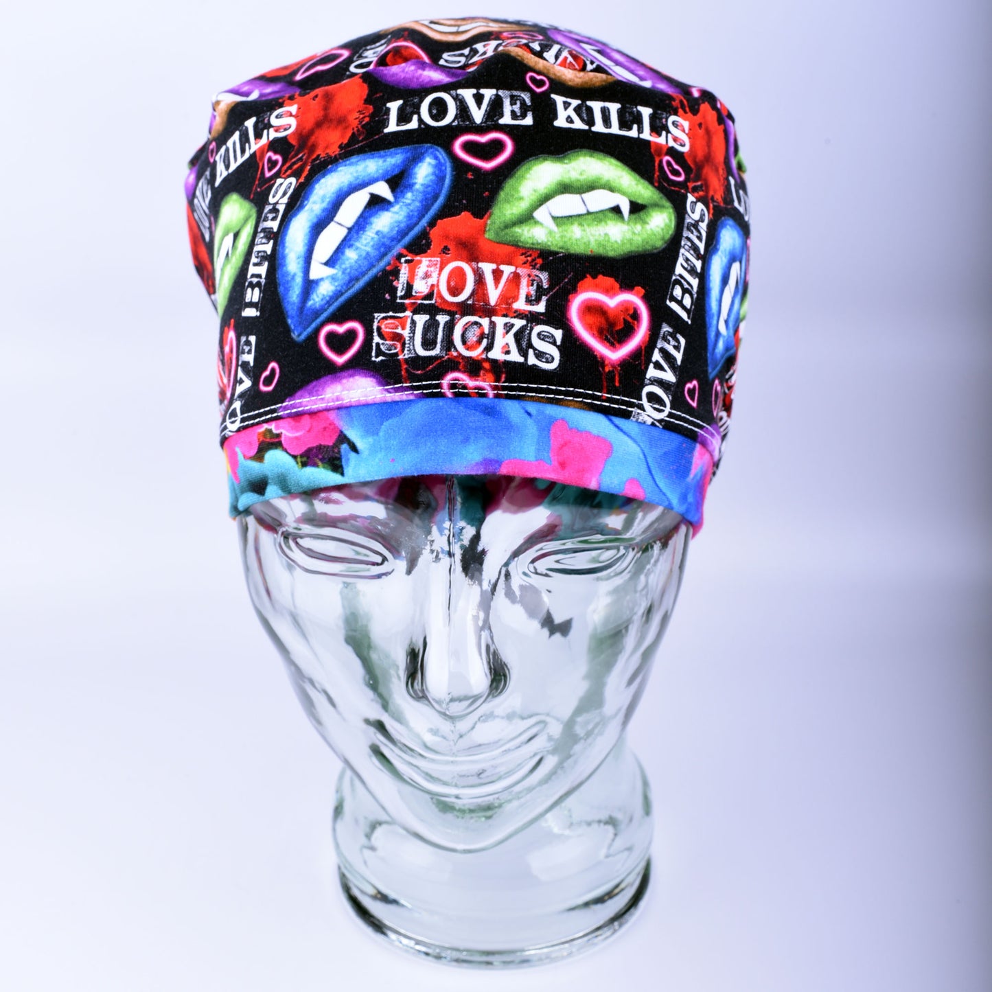 Full-coverage scrub cap featuring an edgy, vibrant Valentine's Day pattern of colorful conversation hearts with phrases like "Love Kills" and "Love Sucks" on a black background, displayed on a clear glass mannequin head.