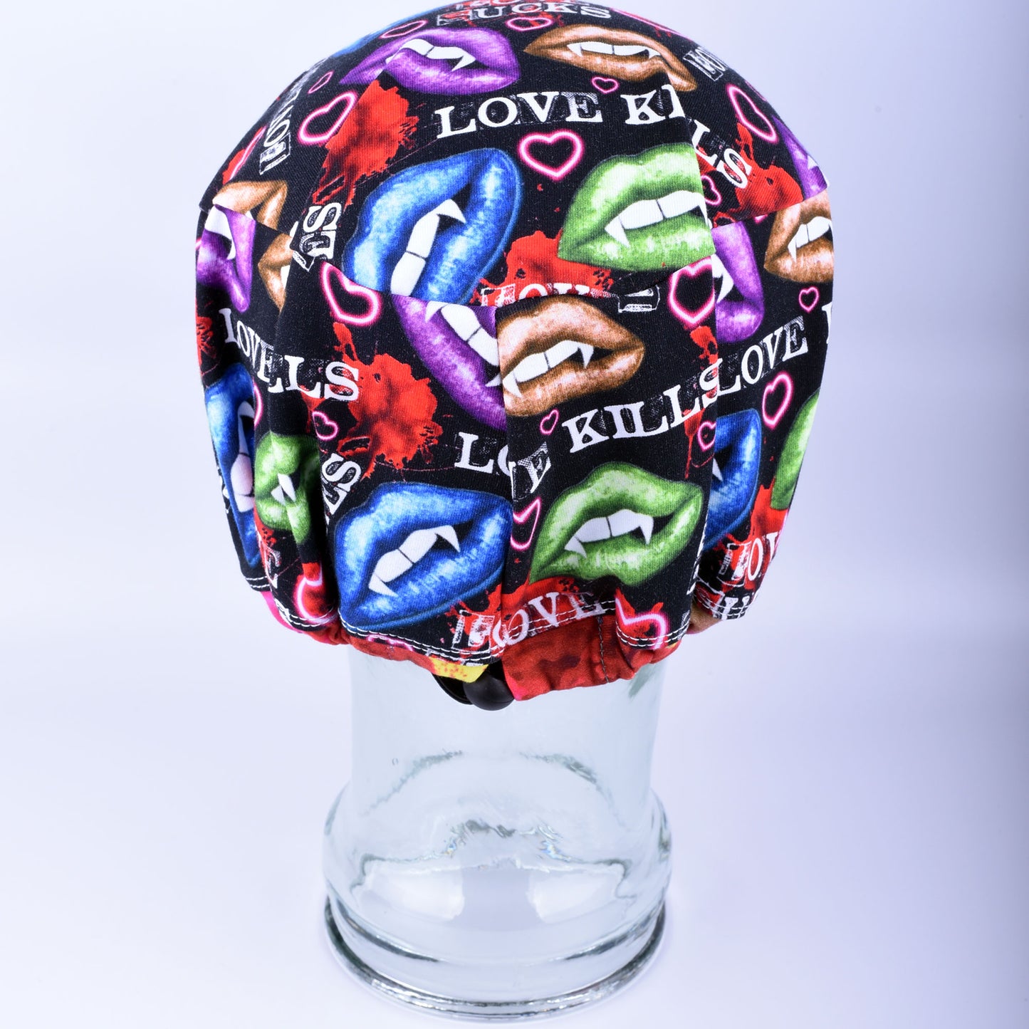 Full-coverage scrub cap featuring an edgy, vibrant Valentine's Day pattern of colorful conversation hearts with phrases like "Love Kills" and "Love Sucks" on a black background, displayed on a clear glass mannequin head.
