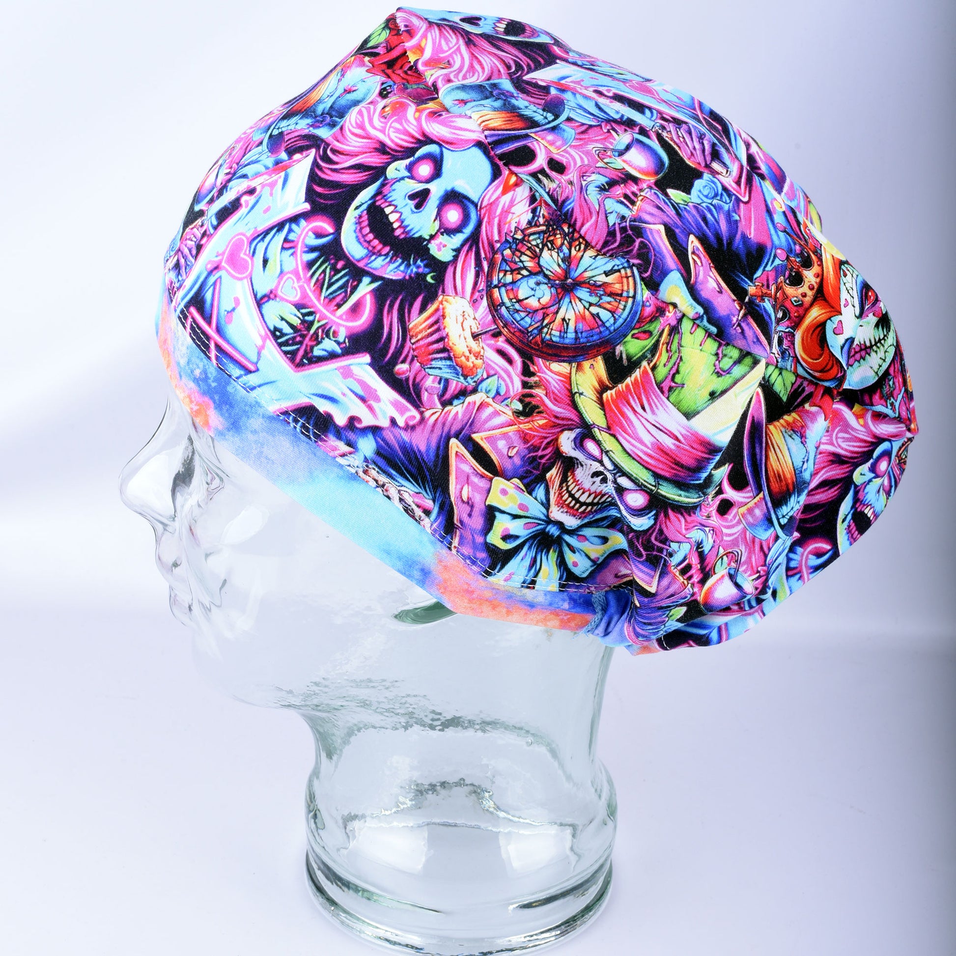 A multicolor scrubs cap with a vibrant, detailed graphic print featuring skulls, flowers, and abstract designs in pink, blue, purple, and green, displayed on a clear glass mannequin head against a light gray background.