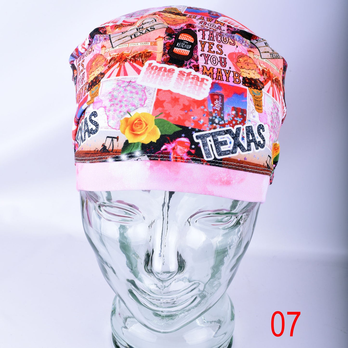 A multicolor Texas-themed scrub cap with a collage pattern featuring 'TEXAS' text, a rose, and food imagery, displayed on a clear glass mannequin head against a light gray background.