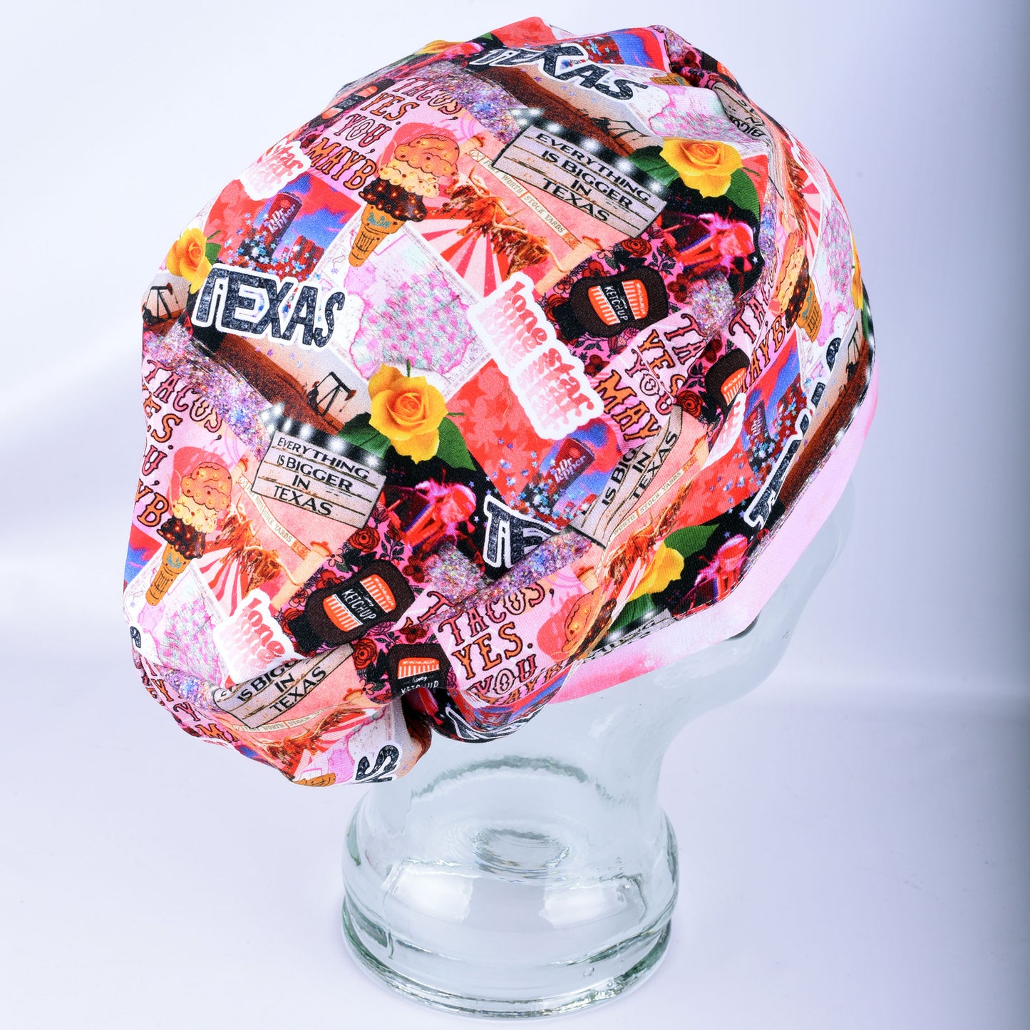 A multicolor scrub cap featuring a collage print of Texas-themed stickers, signs, and illustrations, displayed on a clear glass mannequin head against a light gray background.