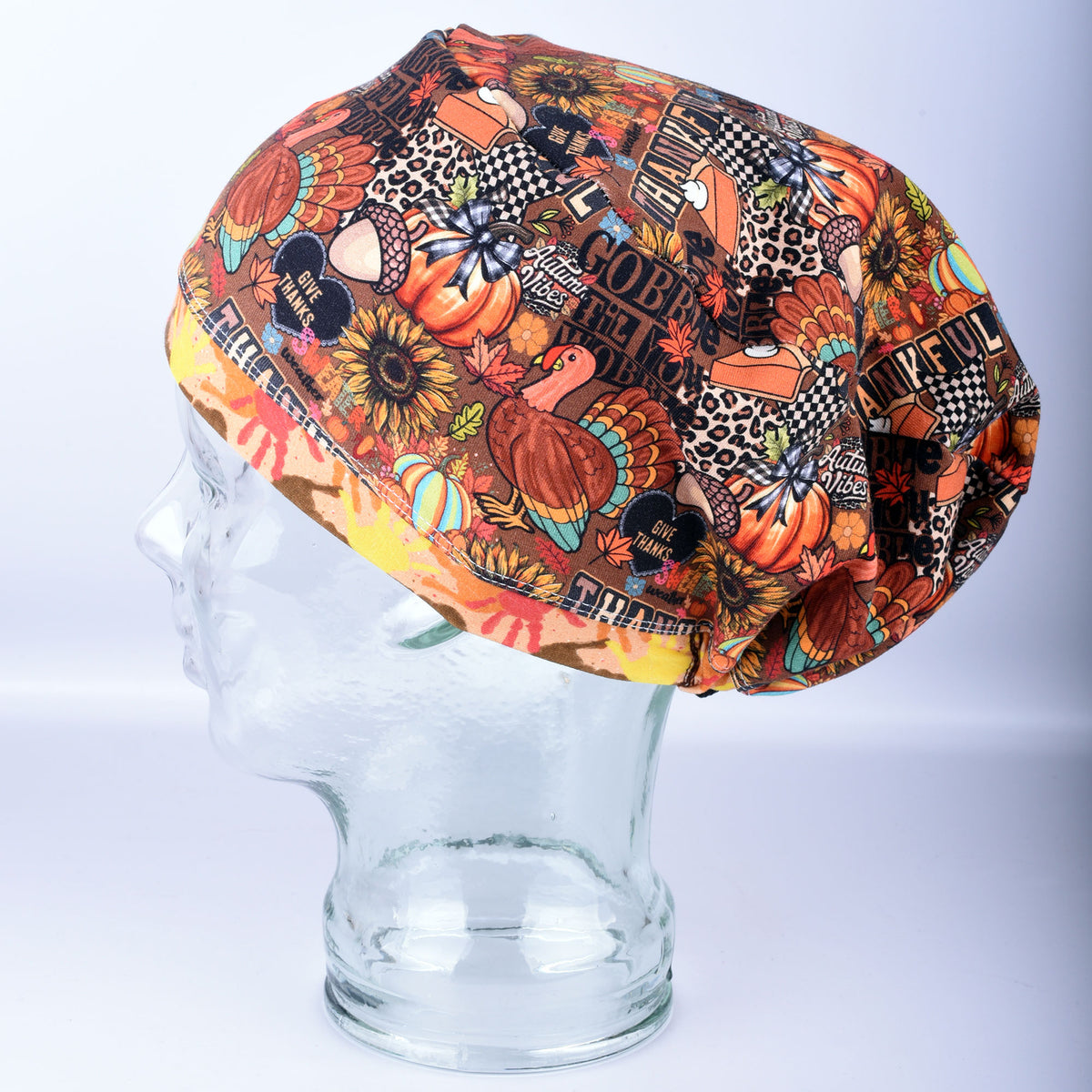 A multicolor Thanksgiving-themed scrub cap with a busy pattern of pumpkins, turkeys, sunflowers, and autumn motifs, displayed on a clear glass mannequin head against a light gray background.