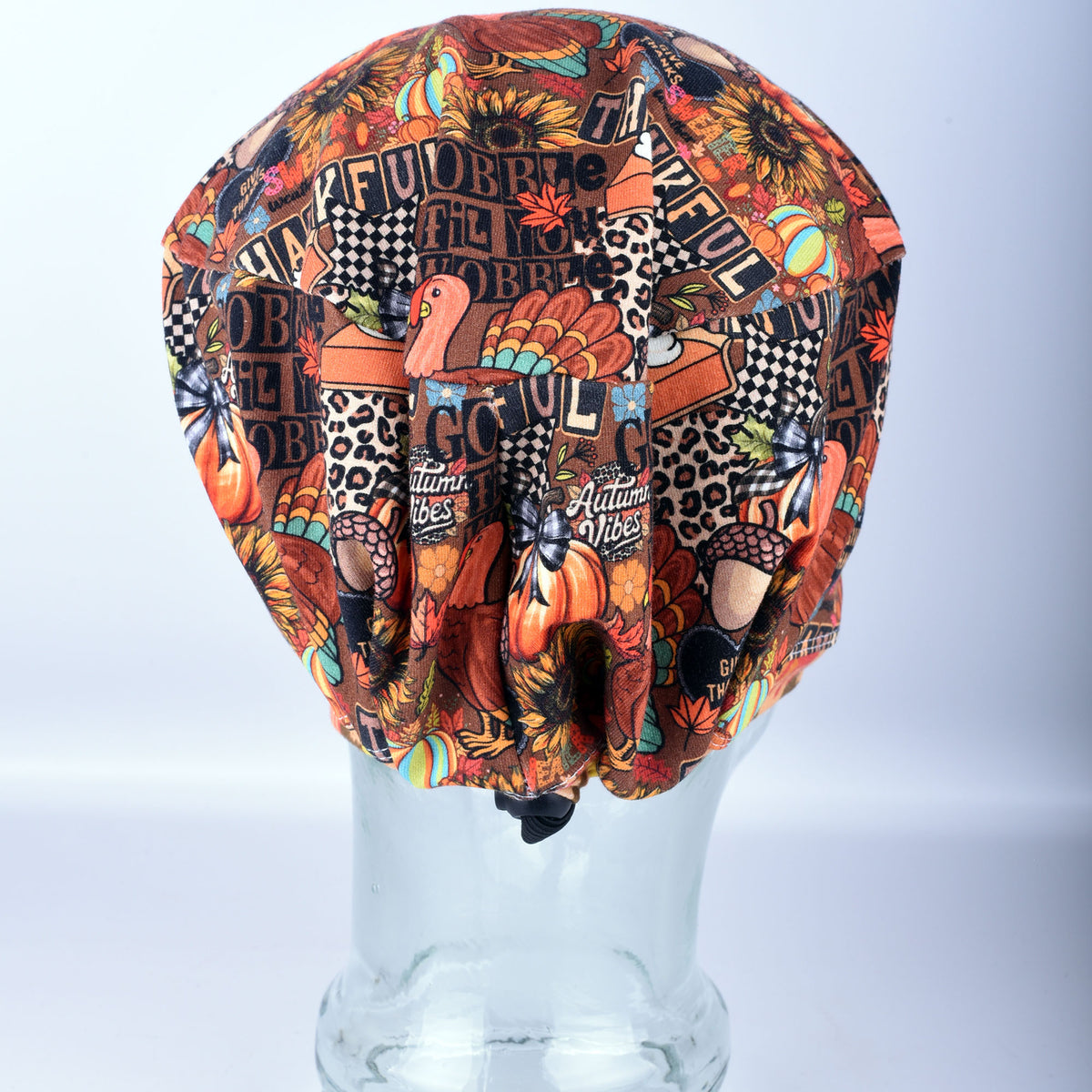 A multicolor scrub cap with a busy autumn-themed collage print featuring pumpkins, turkeys, sunflowers, leopard print, and text, displayed on a clear plastic mannequin head against a light gray background.