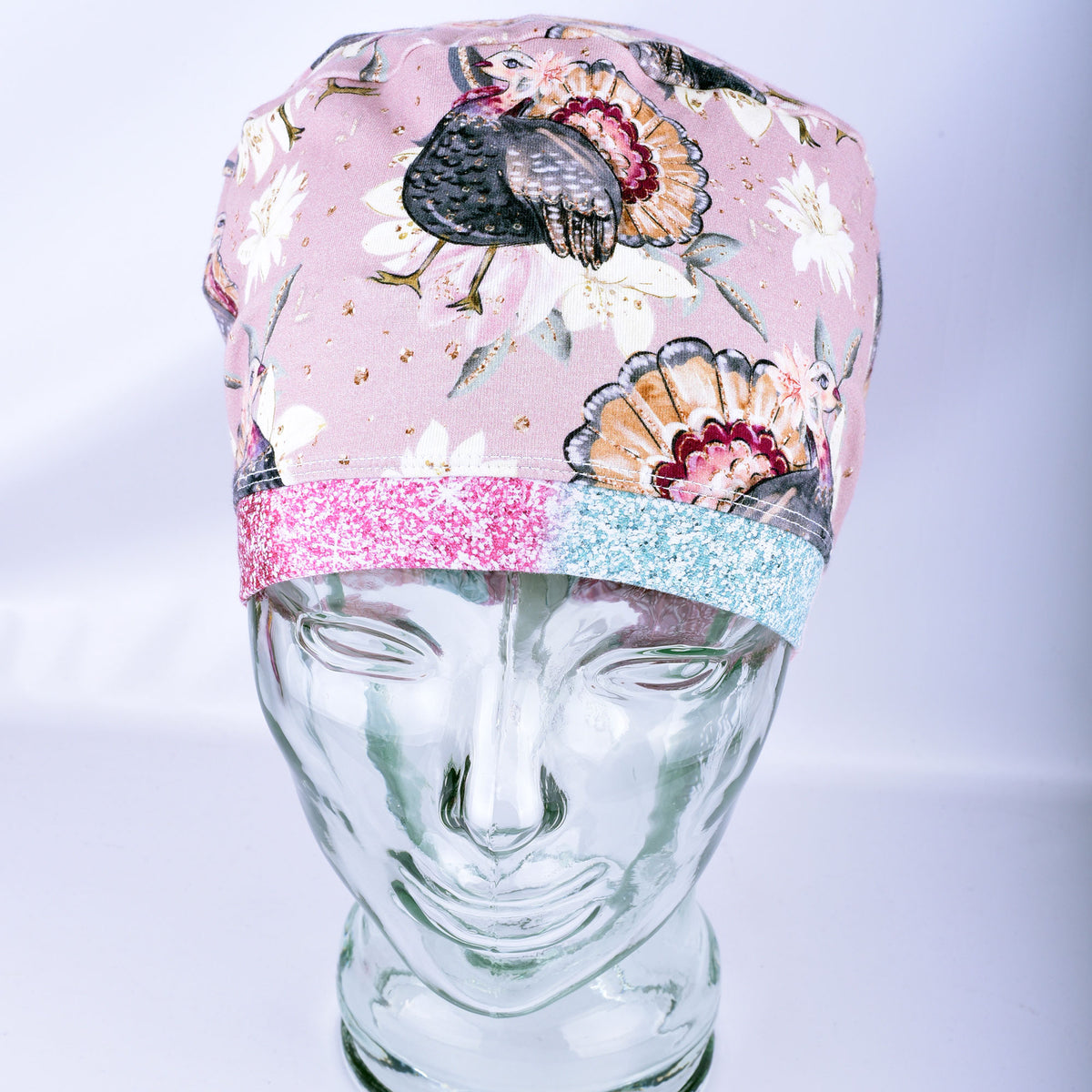 A scrub cap with a turkey and floral print, featuring a glittery pink and light blue split band, displayed on a clear glass mannequin head.