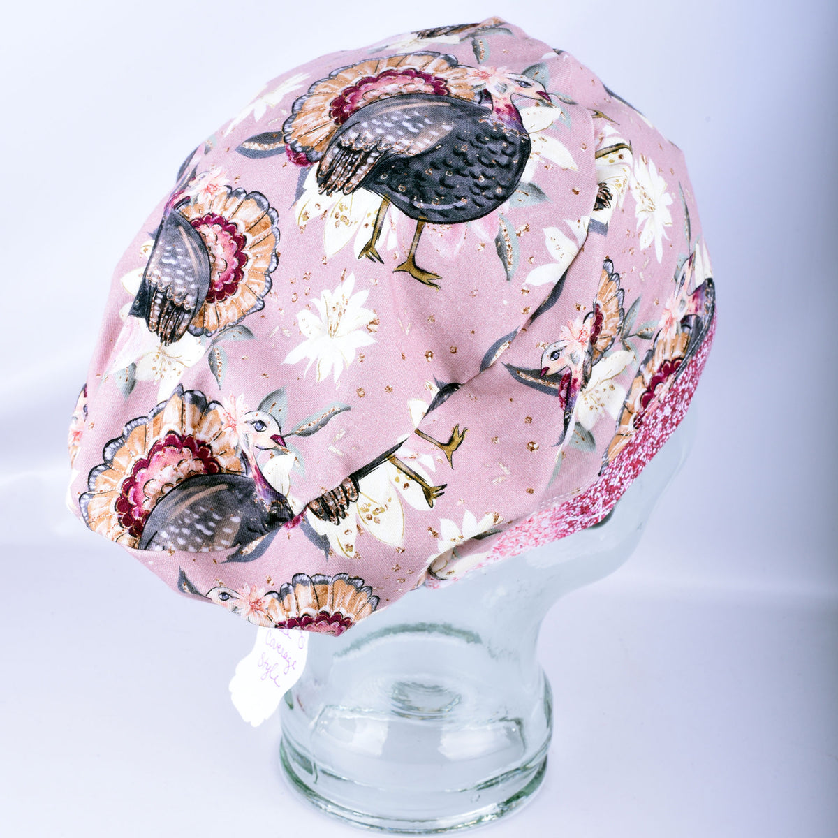 A scrub cap featuring a detailed turkey and floral print, displayed on a clear glass mannequin head against a light gray background.