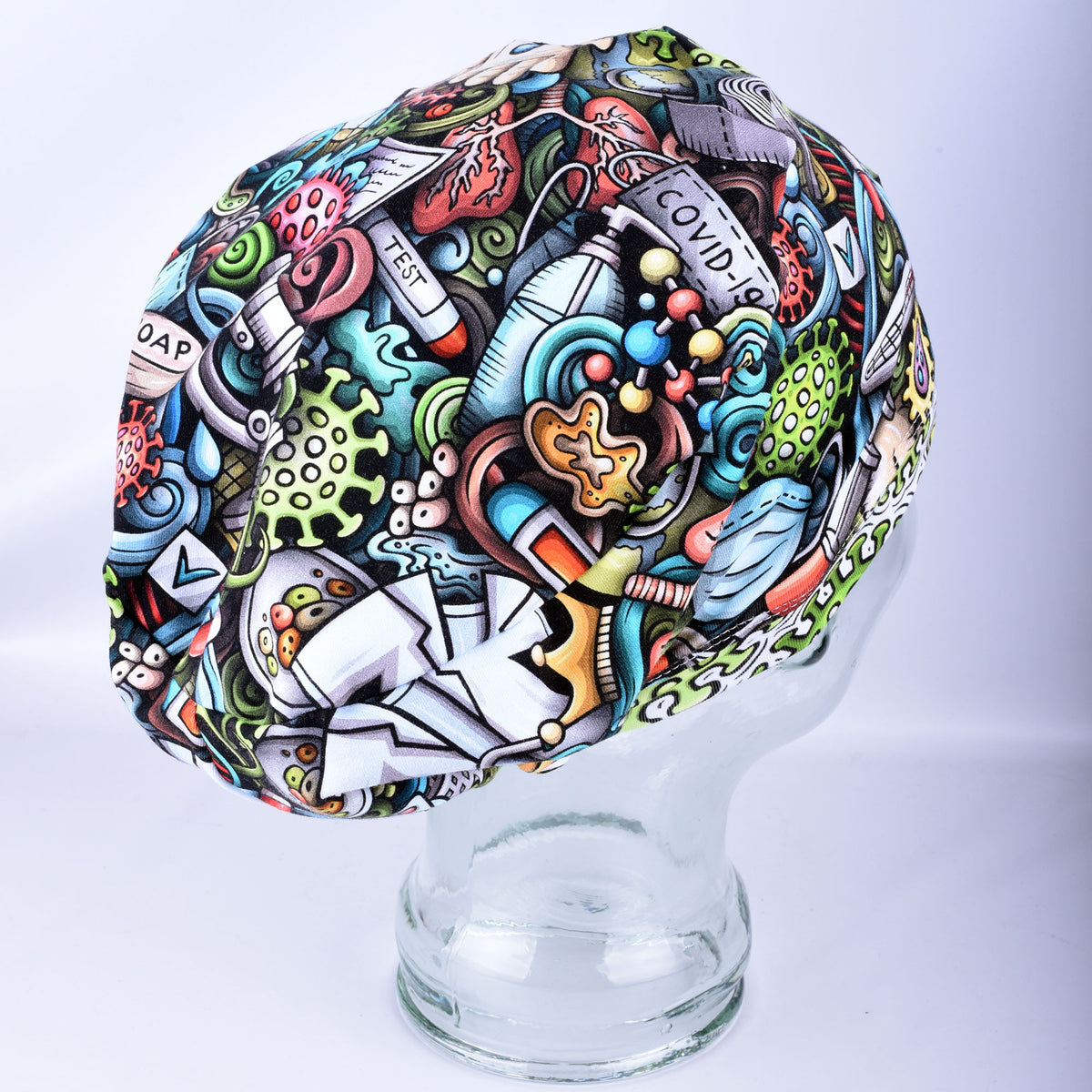 A multicolor medical scrub cap with a detailed cartoon-style graphic print featuring medical icons, viruses, and the text 'COVID-19', displayed on a clear glass mannequin head against a light gray background.