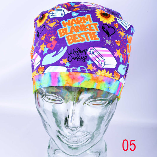 A purple scrub cap with a colorful cartoon print and the words 'WARM BLANKET BESTIE' and 'Warm Cozy' displayed on a clear mannequin head. The cap features illustrations of sunflowers, hearts, and medical-themed icons, with a rainbow tie-dye style band.