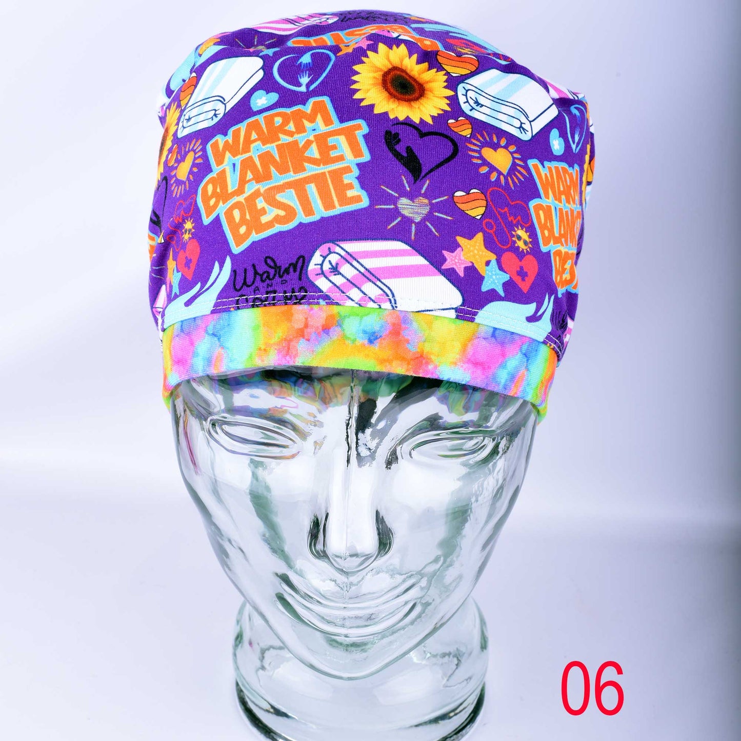 A purple scrub cap with a colorful graphic print featuring the words 'WARM BLANKET BESTIE' and illustrations, displayed on a clear mannequin head against a light gray background.