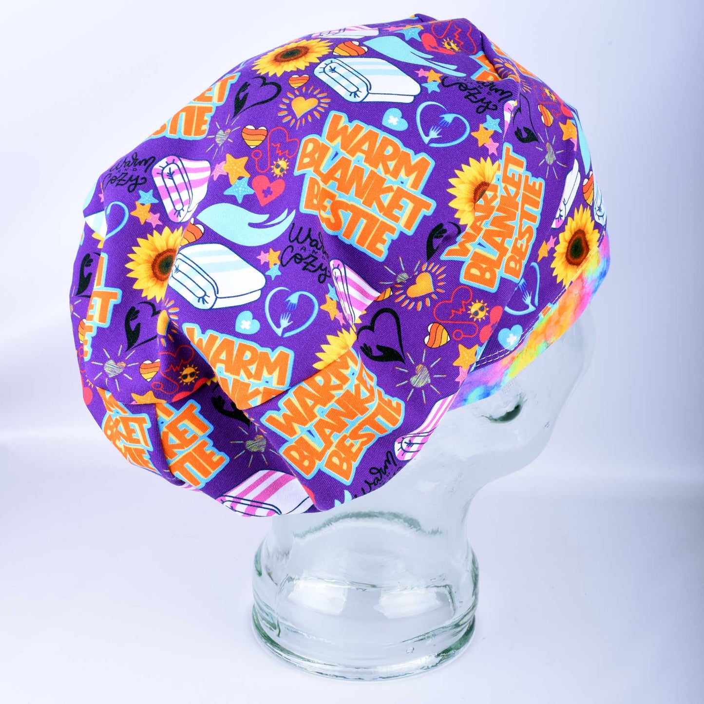A purple medical scrub cap with a colorful 'Warm Blanket Bestie' cartoon print, displayed on a clear glass mannequin head against a white studio background.