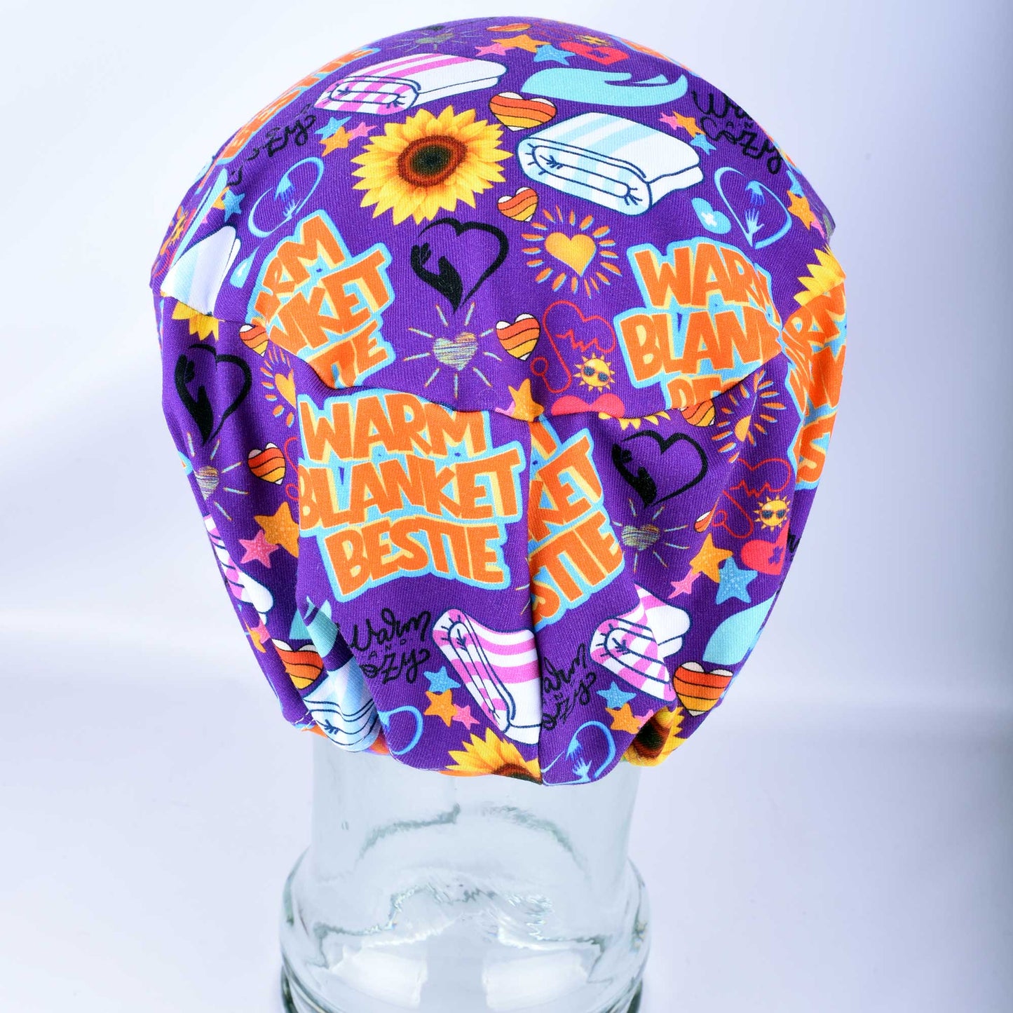 Back view of a purple scrub cap with a colorful 'WARM BLANKET BESTIE' graphic print, displayed on a clear glass mannequin head against a light gray background.