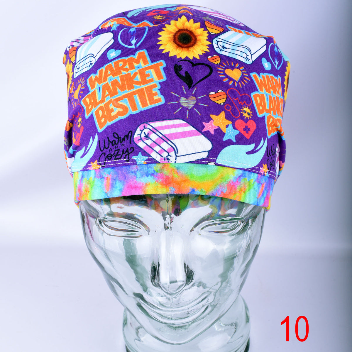 A purple scrub cap with a colorful cartoon print and the words 'WARM BLANKET BESTIE' and 'Warm Cozy' displayed on a clear mannequin head. The cap features illustrations of sunflowers, hearts, and medical-themed icons, with a rainbow tie-dye style band.