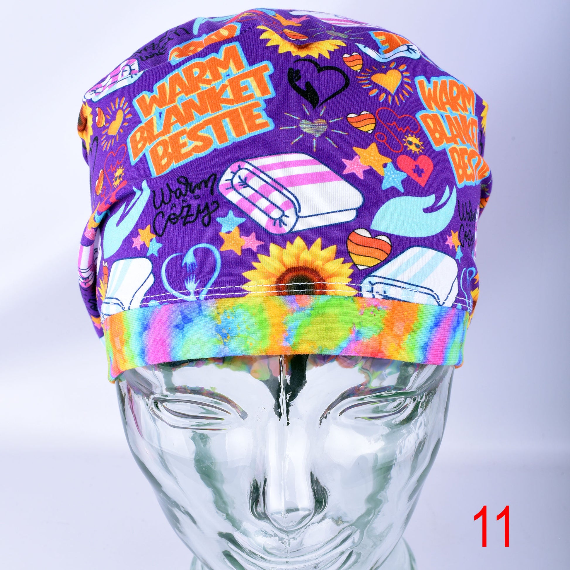 A purple fabric scrub cap with a colorful cartoon print featuring the text 'WARM BLANKET BESTIE' and illustrations of a sunflower, folded blankets, and hearts, displayed on a clear mannequin head.