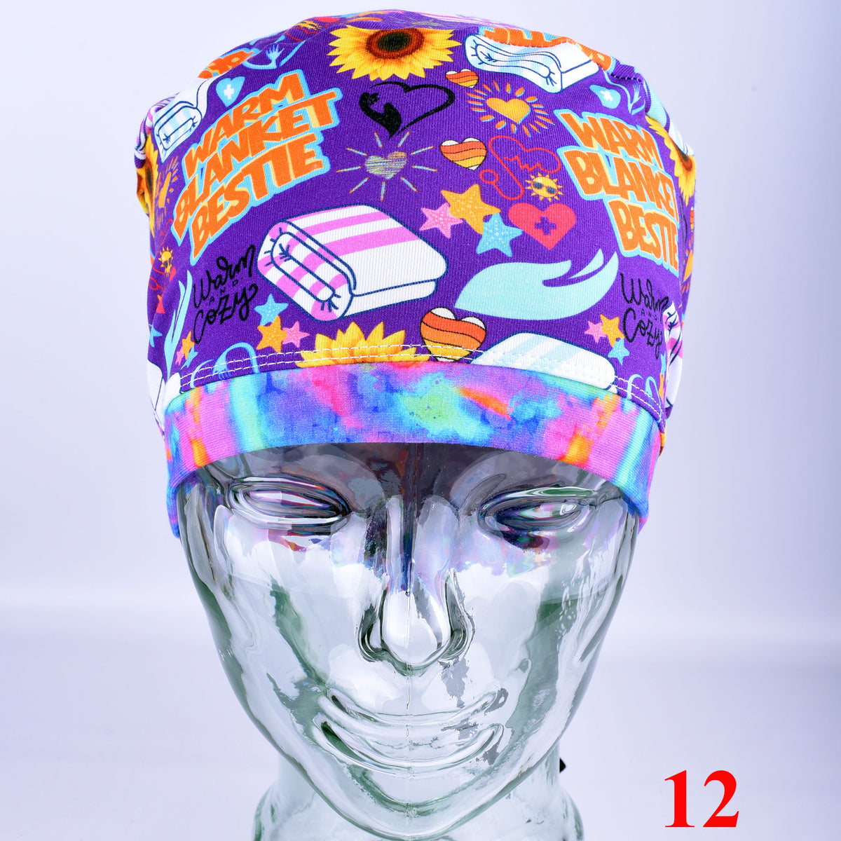 A purple fabric scrub cap with a colorful cartoon print featuring the text 'WARM BLANKET BESTIE' and illustrations of a sunflower, folded blankets, and hearts, displayed on a clear mannequin head.