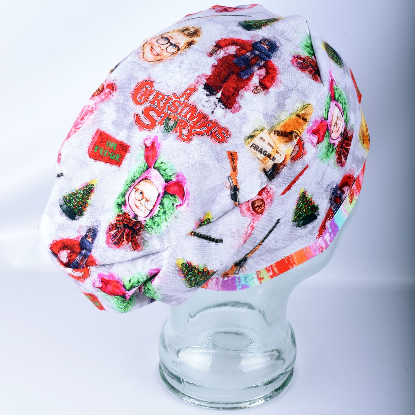 A fabric scrub cap featuring a colorful Christmas-themed print with illustrations of Santa Claus and Christmas trees, displayed on a clear glass mannequin head against a light gray background.