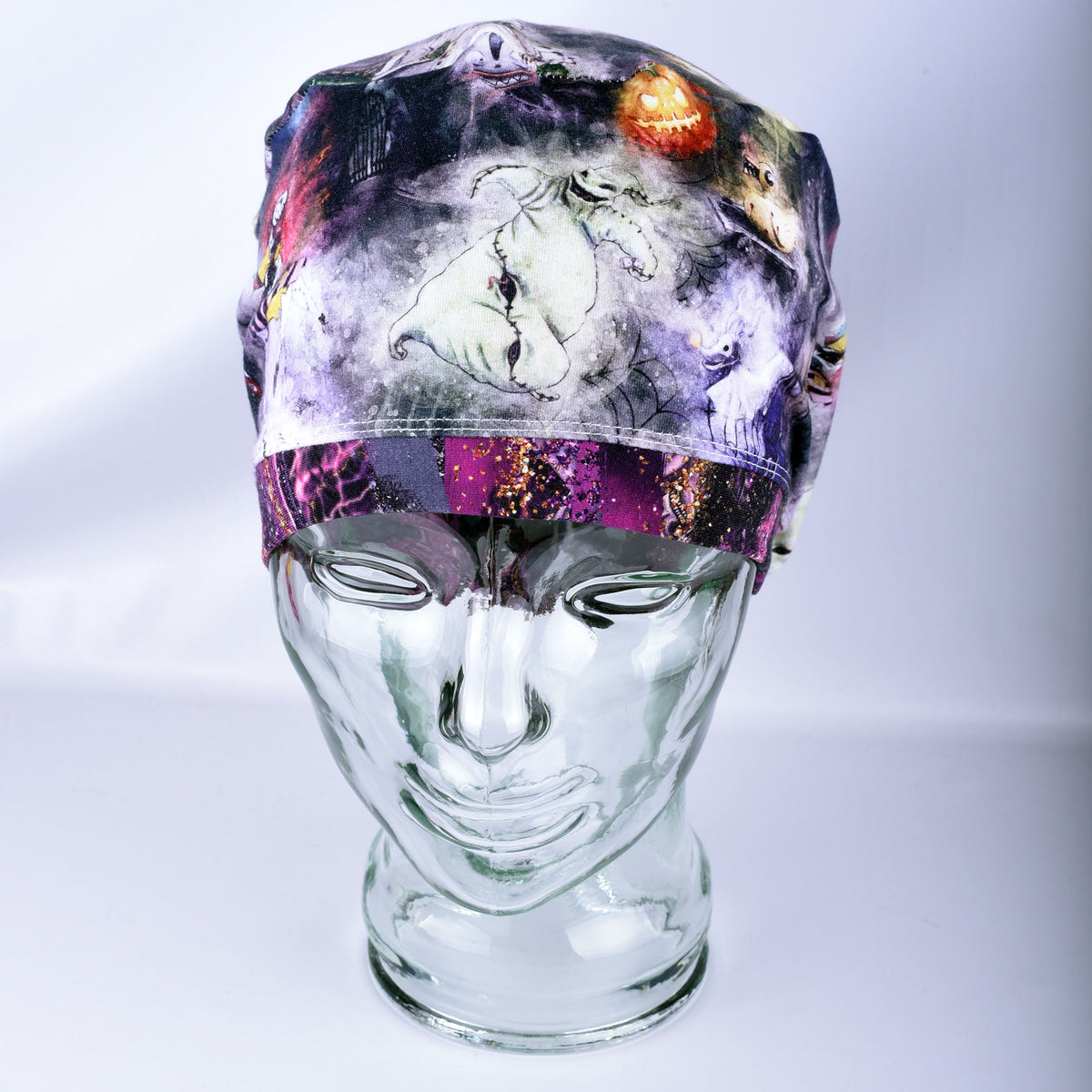 A Halloween-themed surgical scrub cap displayed on a clear glass mannequin head. The cap features a dark, spooky print with a white ghost, a jack-o'-lantern, and purple glittery accents.