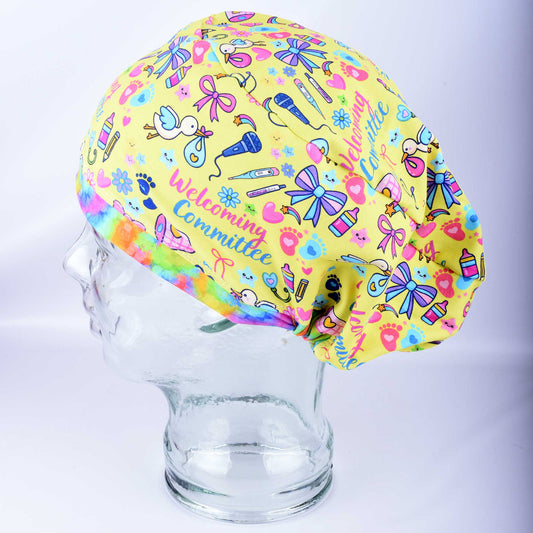 Side view of the Welcoming Committee Euro-style scrub cap on a clear mannequin head, showing the vibrant yellow, pink, and blue infant-themed pattern and comfortable full-coverage fit.