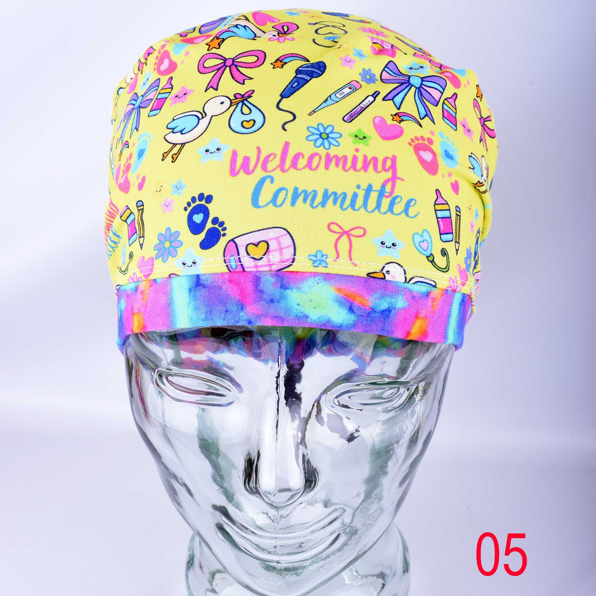 A full-coverage Euro-style medical scrub cap with a colorful baby-themed print displayed on a clear mannequin head. The white fabric features a vibrant pattern of baby bottles, safety pins, and toys with the cursive text "Welcoming Committee" printed across the front.
