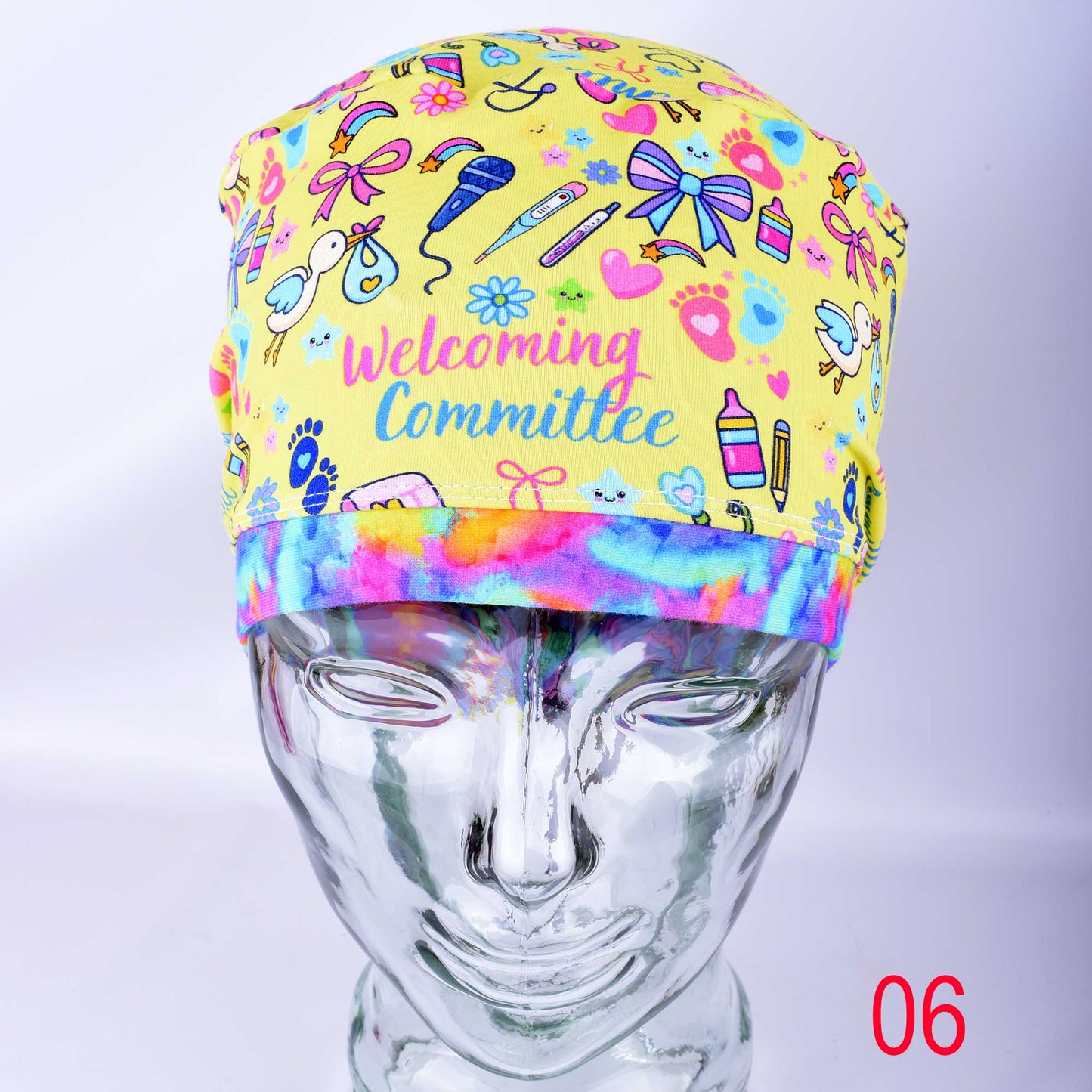 A full-coverage Euro-style medical scrub cap with a colorful baby-themed print displayed on a clear mannequin head. The white fabric features a vibrant pattern of baby bottles, safety pins, and toys with the cursive text "Welcoming Committee" printed across the front.