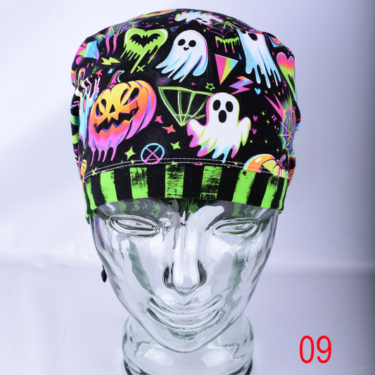 A black scrub hat with a vibrant neon Halloween print featuring ghosts, pumpkins, and hearts, displayed on a clear glass mannequin head against a light gray background.