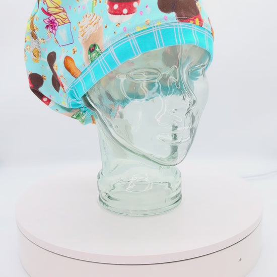 A 14-second rotating product video showing a 360-degree view of the Magical Snacks Full Coverage Scrub Cap on a clear mannequin head. The medical hat features a vibrant light blue fabric patterned with theme park treats like popcorn buckets, churros, soft pretzels, hot dogs, and mouse-shaped ice cream bars, finished with a contrasting blue-and-white striped brim.